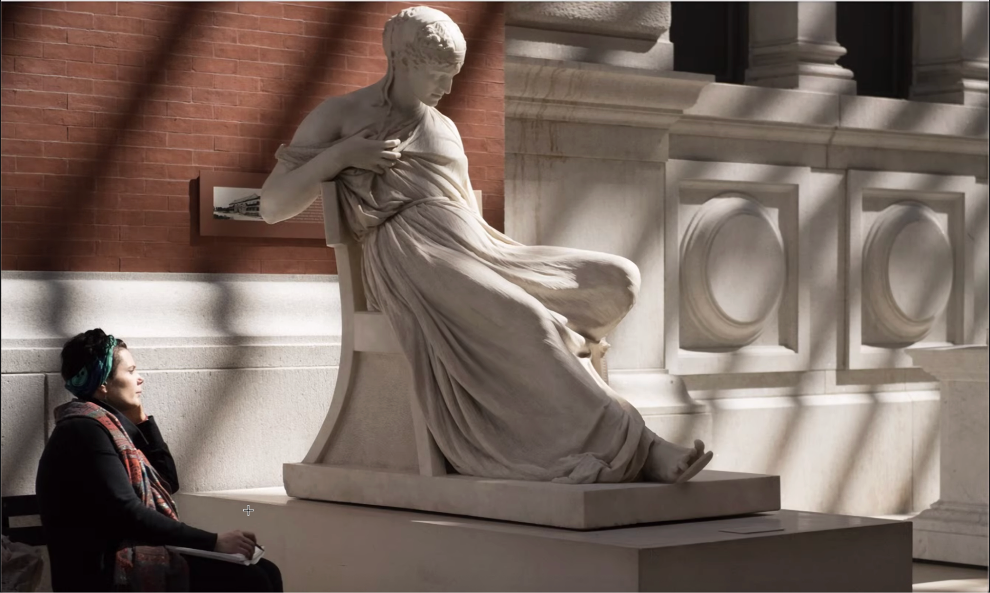 A woman sits on a bench facing a marble sculpture of a figure draped in flowing clothing, with their head bowed. The setting includes warm brick walls and soft shadows, enhancing the tranquil atmosphere of the museum. The woman is taking notes or sketching, wearing headphones and a colorful scarf.