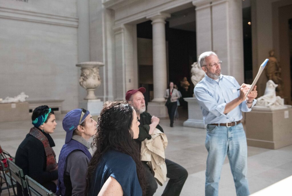 An instructor demonstrates drawing techniques on a sketchbook to a seated group during a museum gallery class surrounded by classical sculptures and architectural columns. Participants observe the demonstration while holding sketchbooks in a spacious gallery illuminated by natural light.