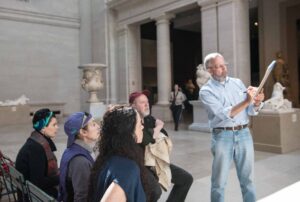 An instructor demonstrates drawing techniques on a sketchbook to a seated group during a museum gallery class surrounded by classical sculptures and architectural columns. Participants observe the demonstration while holding sketchbooks in a spacious gallery illuminated by natural light.