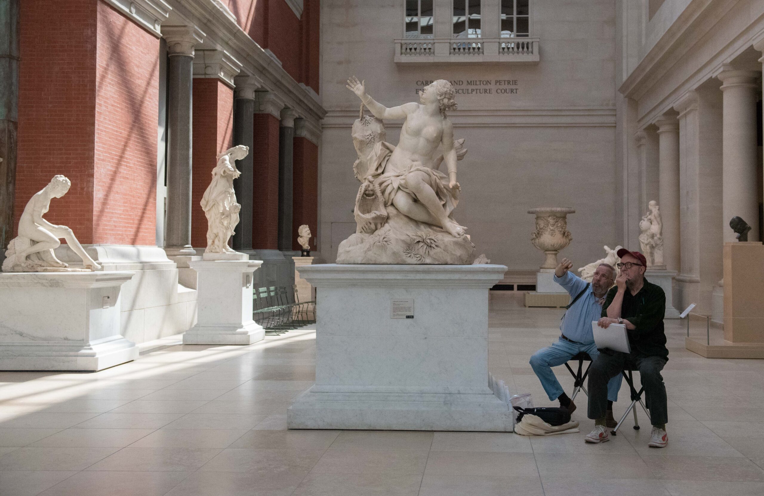 Two men are seated on folding chairs in a museum sculpture court, observing a large marble statue of a figure with outstretched arms, seated on a rock surrounded by foliage. One man gestures towards the statue while the other thoughtfully looks on, holding a sketchbook. Several other sculptures and decorative objects are visible throughout the spacious room, characterized by brick and marble architecture and natural lighting.