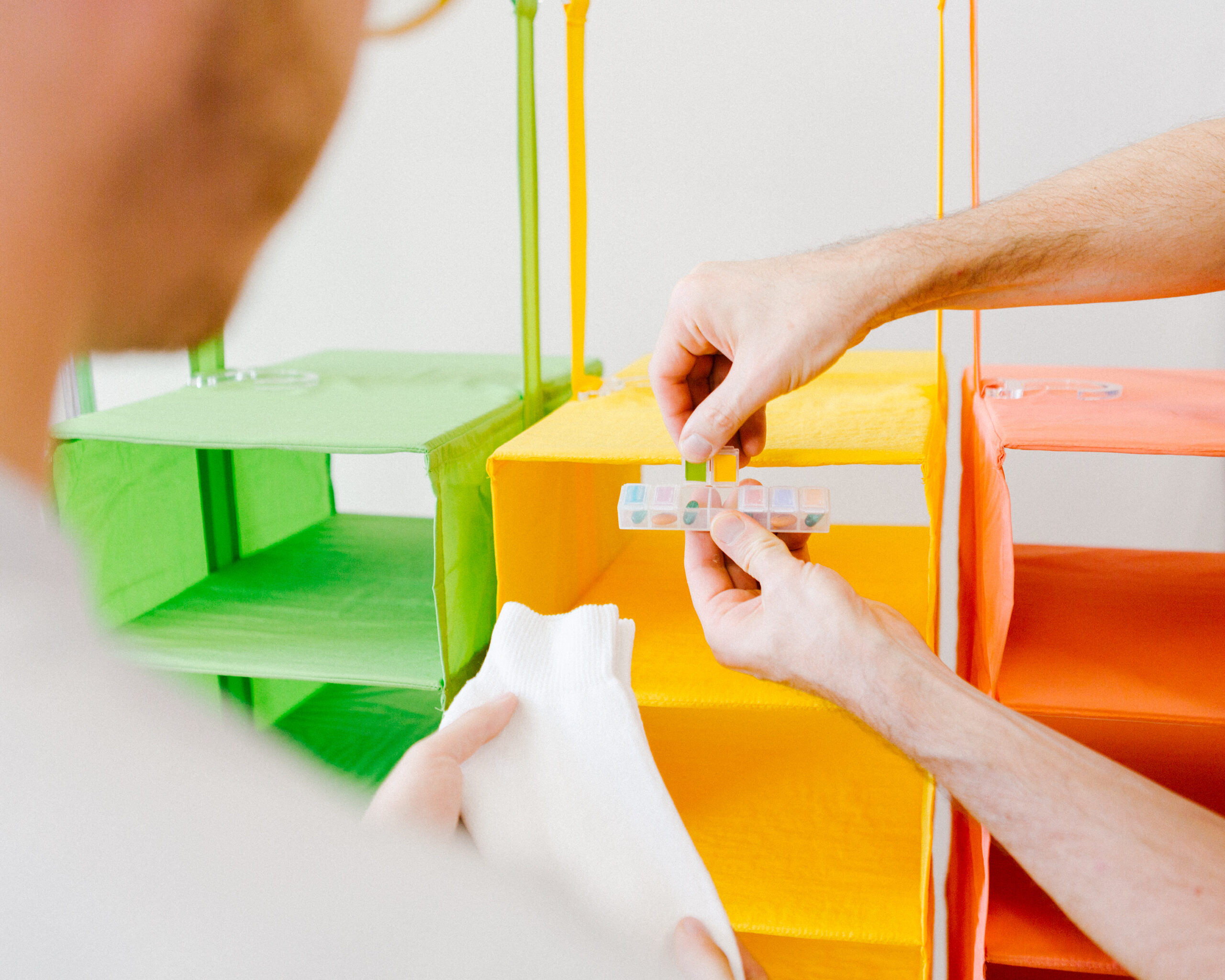 Photo of a pair of hands and the back of a person as they observe and hold a ruler and a pair of cloth which they are holding in front of colored bins.