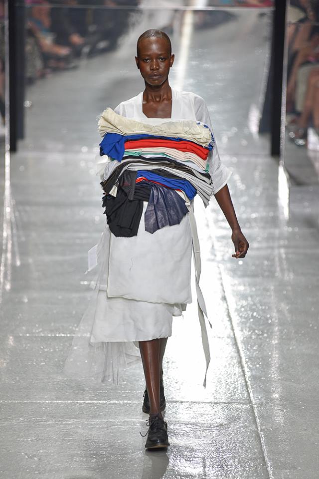 Model seen walking on a runway in a white dress with multicolored fabric on the top half of the dress.
