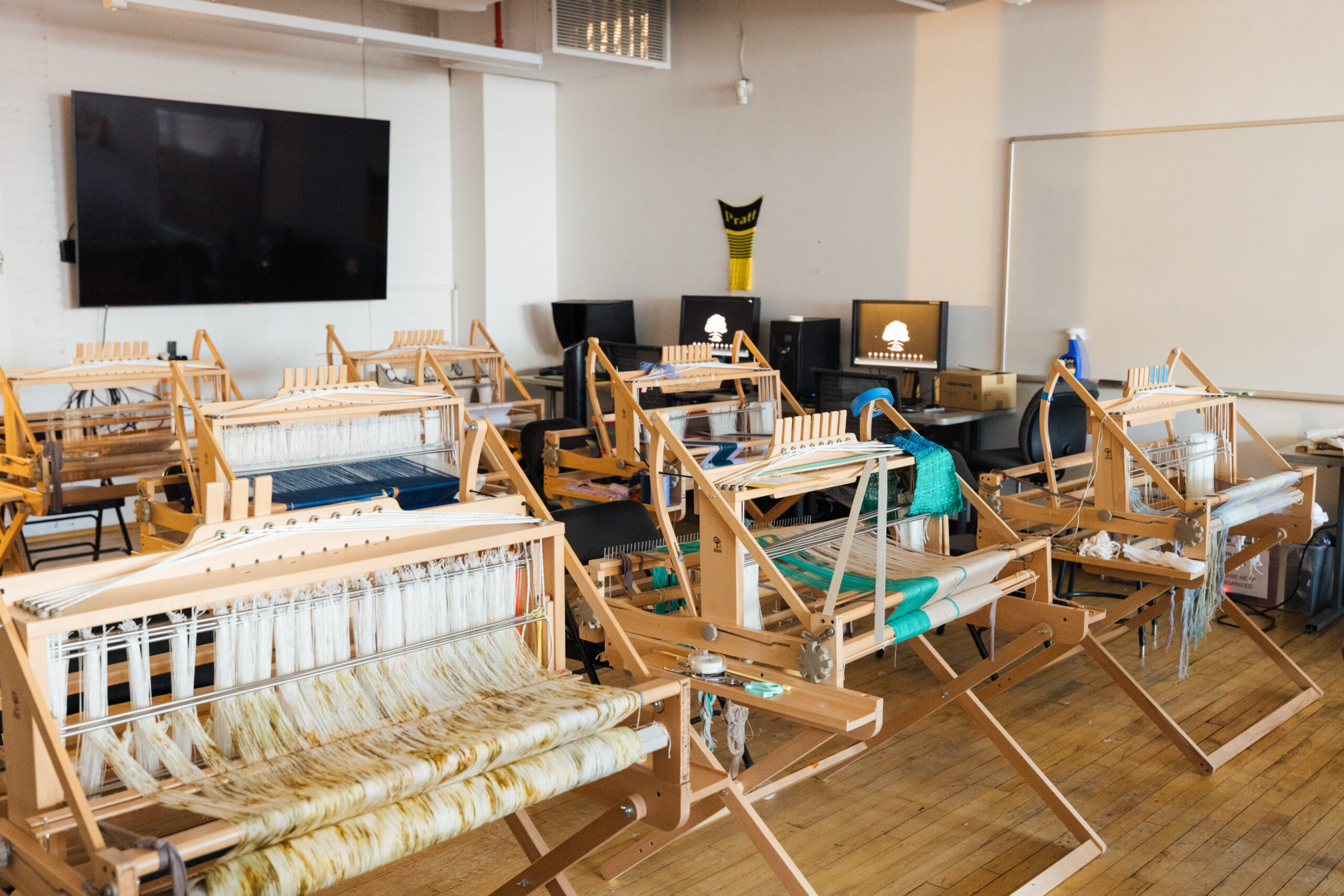 Multiple wooden weaving looms are arranged in rows inside a studio classroom, with partially woven textiles in neutral and green tones stretched across the frames.