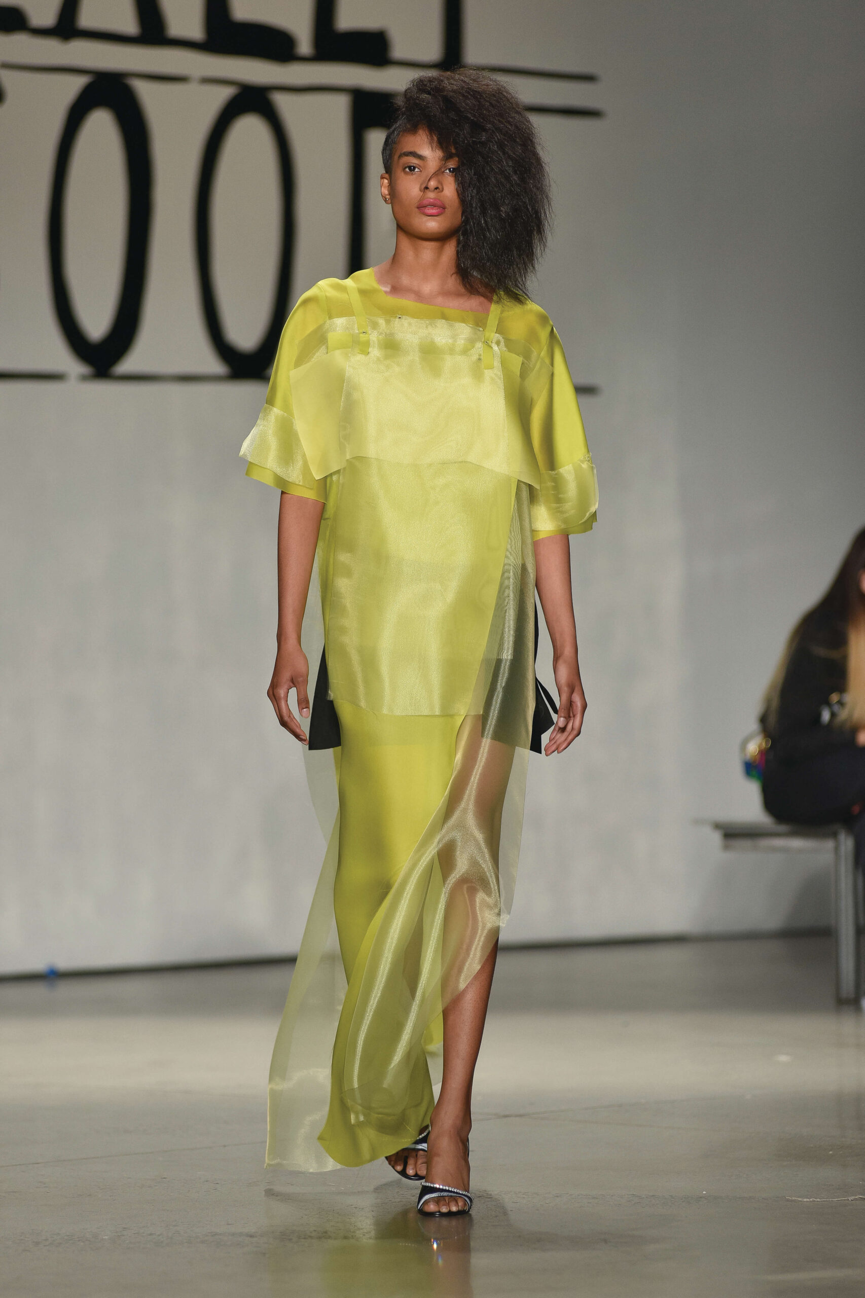 A model walks along a catwalk wearing a long, yellow dress. The bottom of the dress is somewhat translucent.
