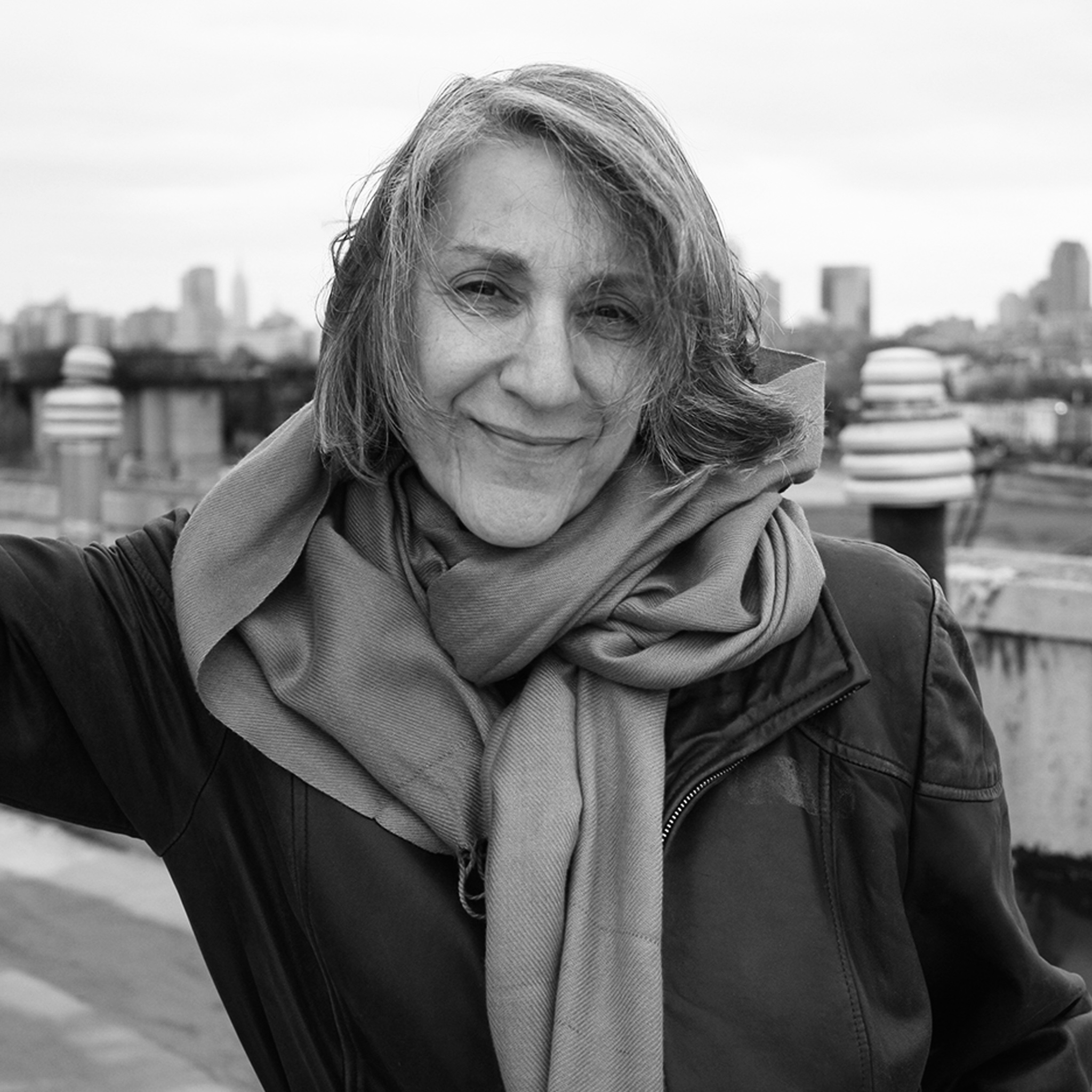 An older woman with shoulder-length gray hair smiles gently while standing outdoors. She is wearing a large scarf and leather jacket. In the background, a city skyline is faintly visible under an overcast sky. The image is in black and white.