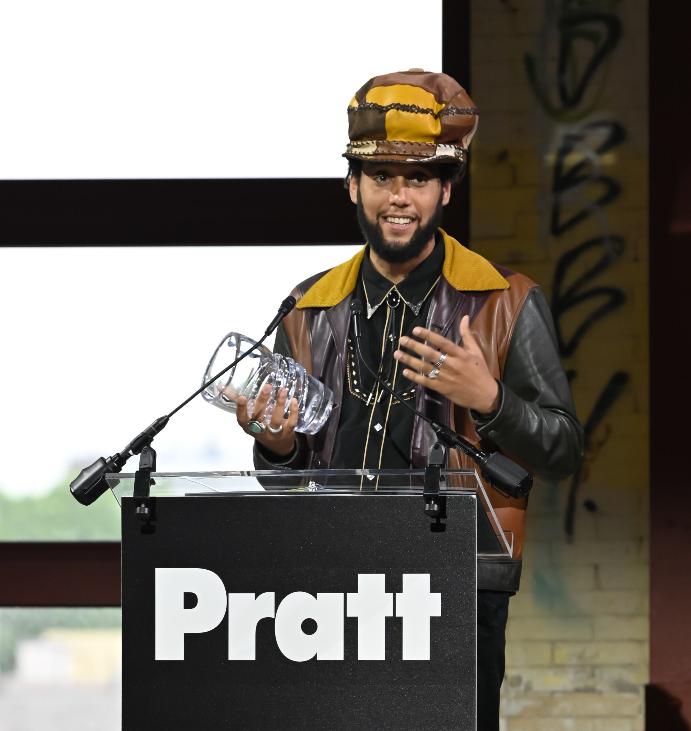 A person stands behind a clear podium with a “Pratt” sign, smiling and speaking while holding an award during a formal fashion-related presentation or ceremony.