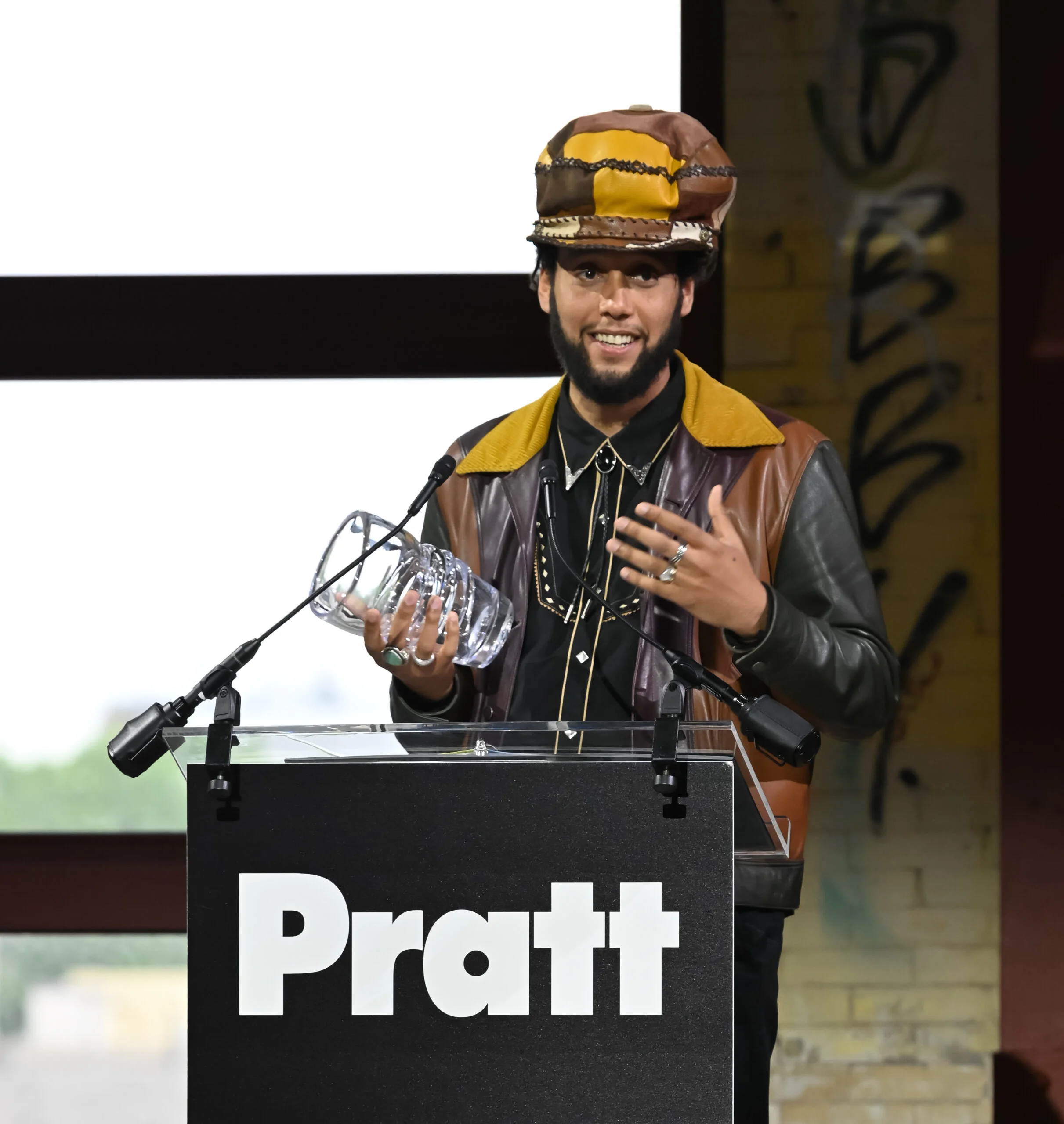 A person stands behind a clear podium with a “Pratt” sign, smiling and speaking while holding an award during a formal fashion-related presentation or ceremony.