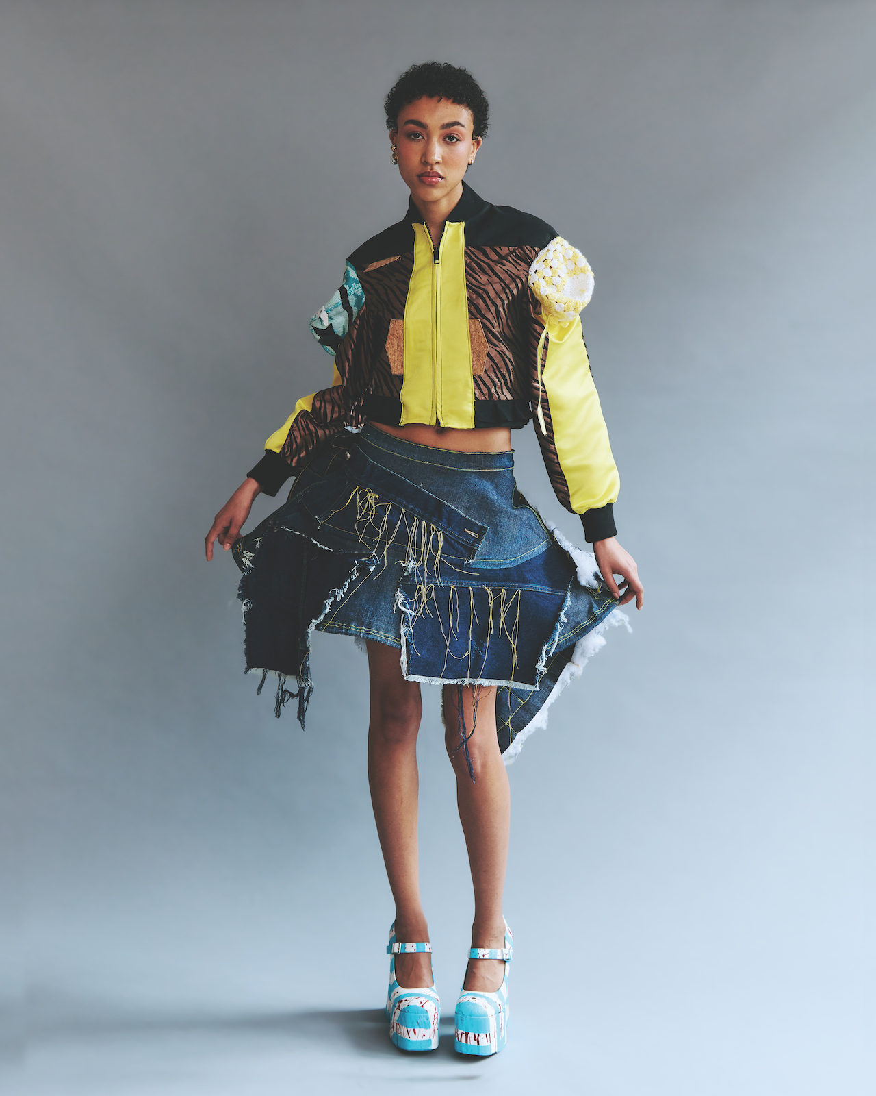 A full-length studio portrait of a model standing against a neutral gray backdrop, wearing a cropped jacket with a bright yellow central panel, patterned brown-and-black fabric, and textured sculptural sleeve details, paired with an asymmetrical deconstructed denim skirt featuring visible stitching, frayed edges, and dangling threads, finished with high platform heels painted in blue, white, and red tones.