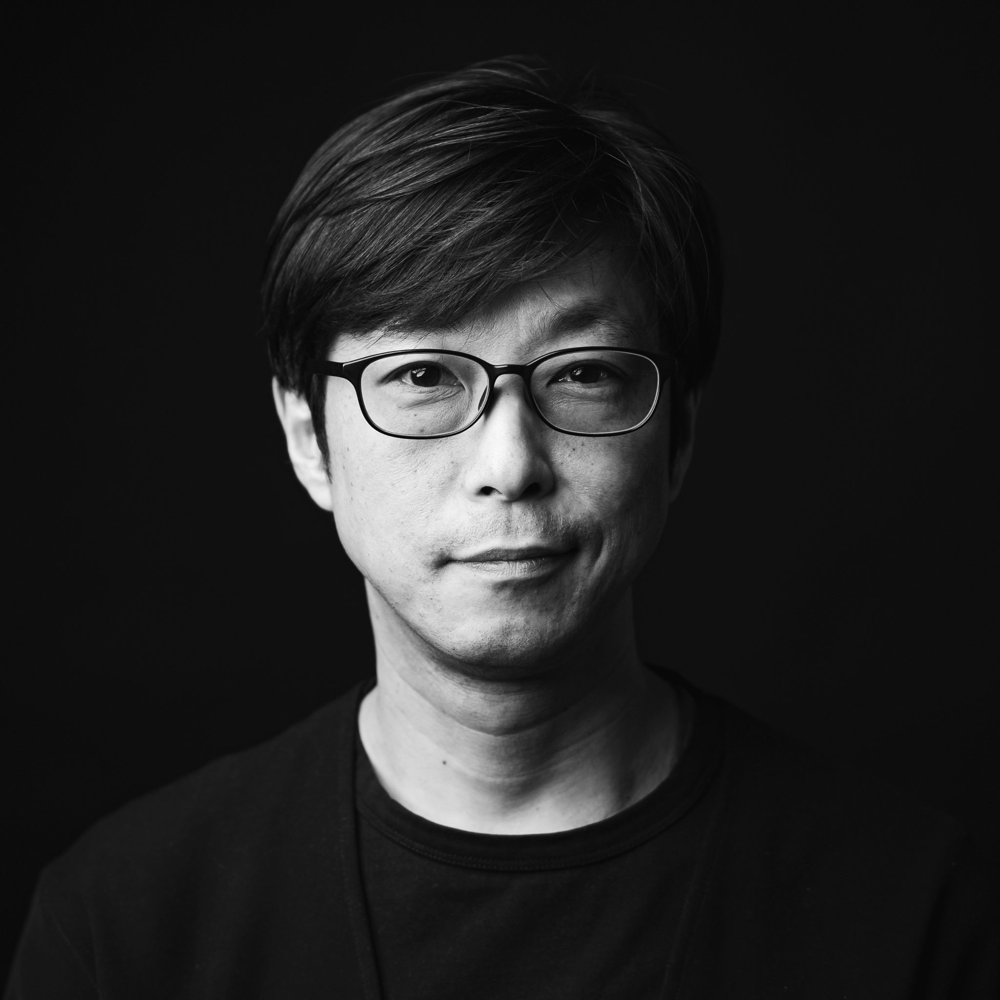 A man with short hair and glasses looks directly into the camera with a calm expression. He wears a dark crew neck shirt. The background is pitch black, drawing focus to the subject’s face in this sharply lit black and white portrait.