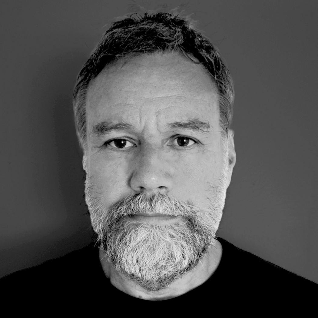 A middle-aged man with short hair and a full gray beard stands against a plain gray background. He wears a black shirt and gazes directly at the camera with a neutral expression. The portrait is in black and white.