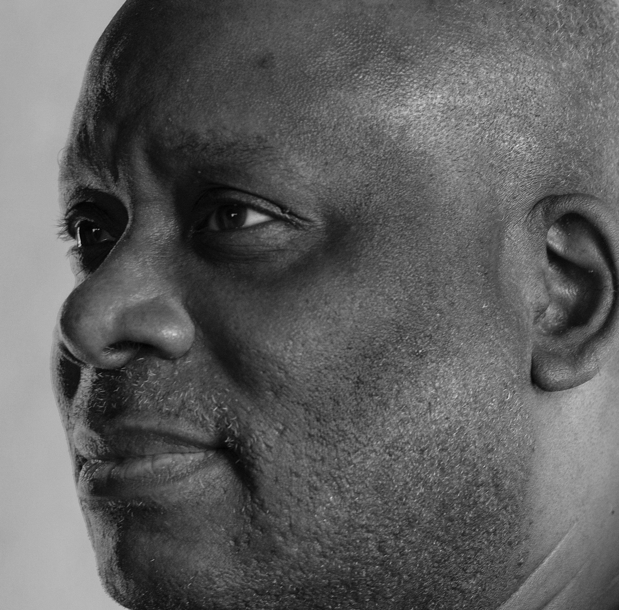 A close-up side profile of a man with short hair and a textured beard, his expression calm and thoughtful. The lighting emphasizes the contours of his face and the fine texture of his skin. The photo is in black and white.