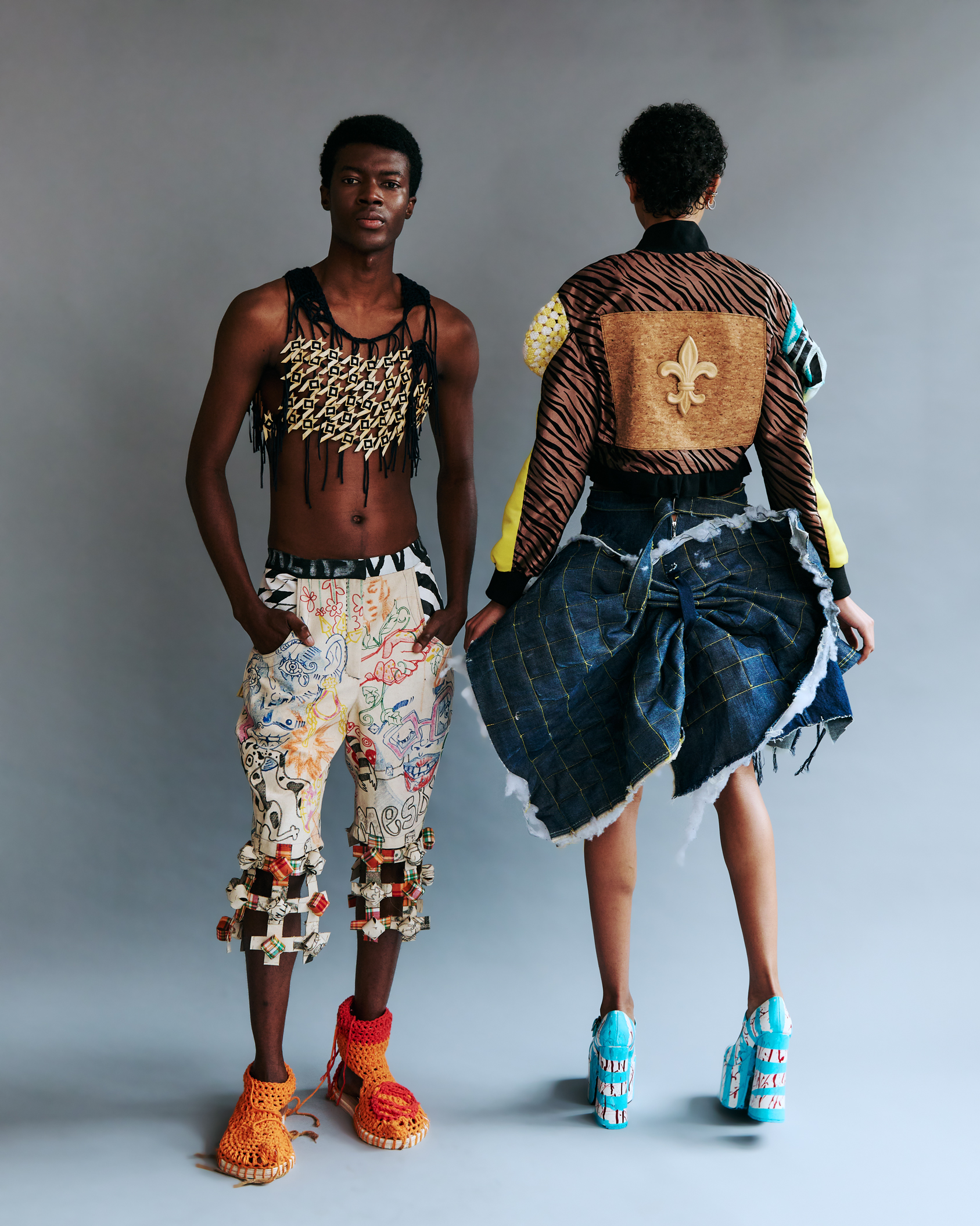 Two fashion models pose against a gray backdrop, one facing forward and one facing away, wearing experimental garments with mixed materials, textures, and bold handcrafted details.