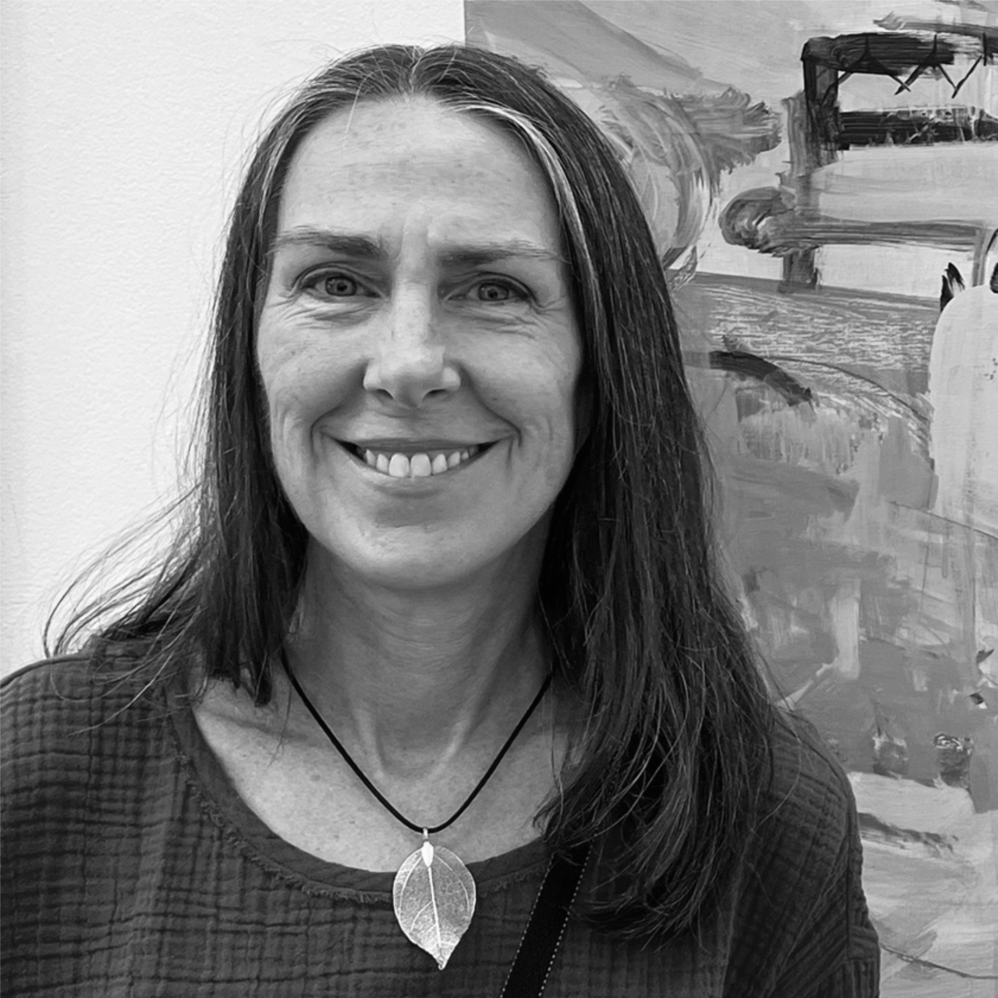 A woman with shoulder-length hair stands indoors, smiling warmly. She wears a floral shirt and hoop earrings, with sunlight streaming through nearby windows. The image has a candid, welcoming feel.