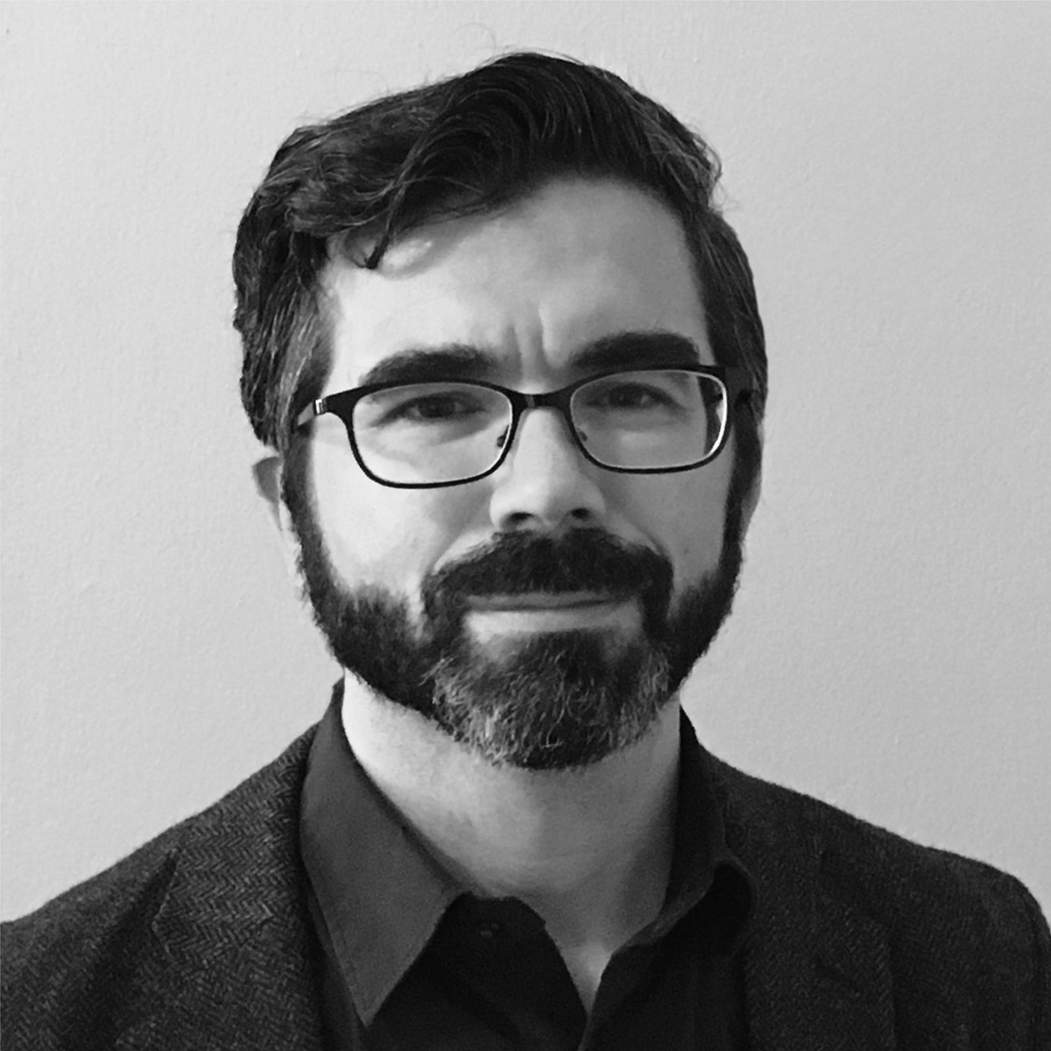A man with a bald head and a thick, graying beard looks out of frame while sitting indoors. He wears large dark-framed glasses and a textured cardigan over a white shirt. The lighting is soft, and the photo is in black and white.