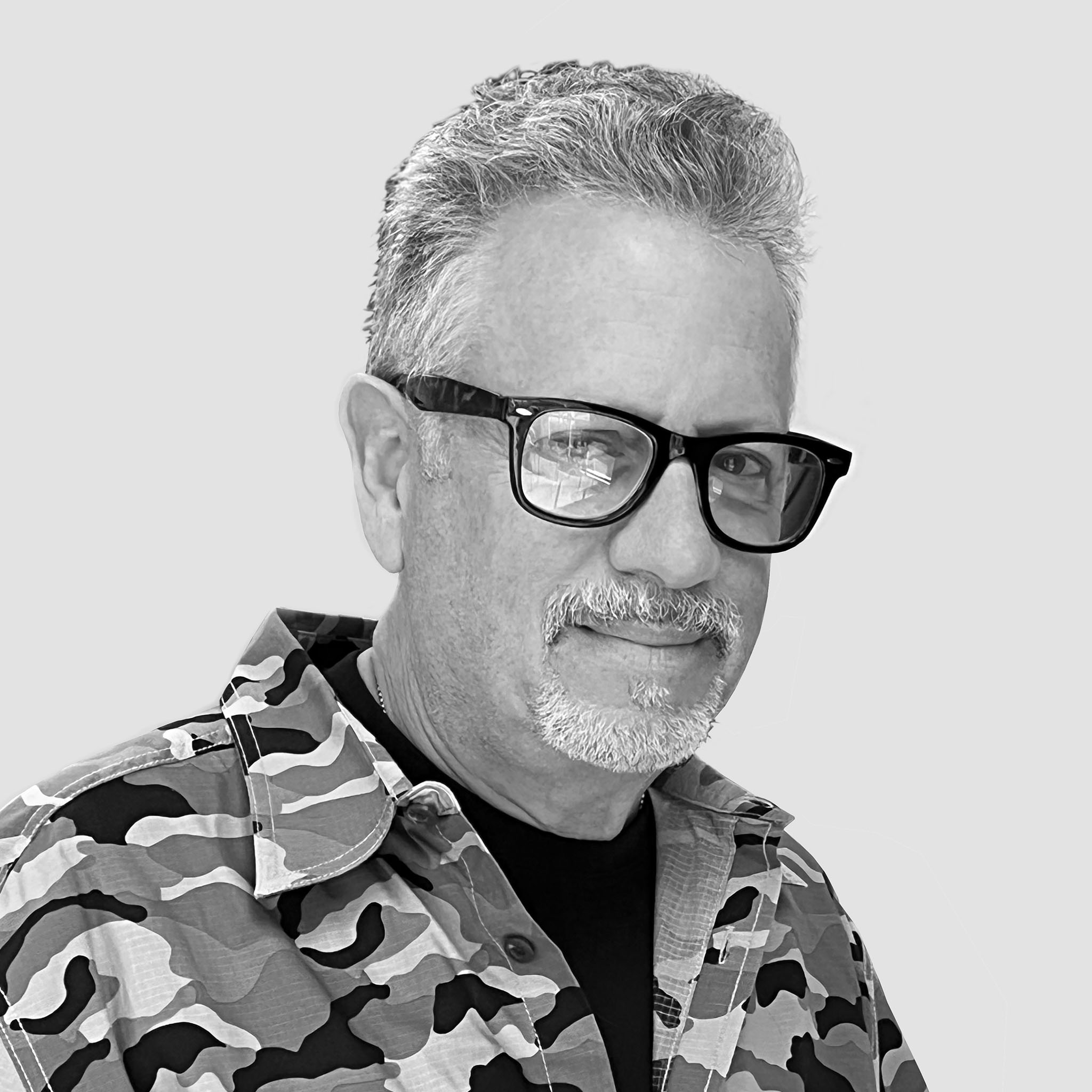 A man with salt-and-pepper hair, a goatee, and black-framed glasses stands against a white background. He wears a camouflage-print shirt and a confident smile. The black and white image feels bold and casual.