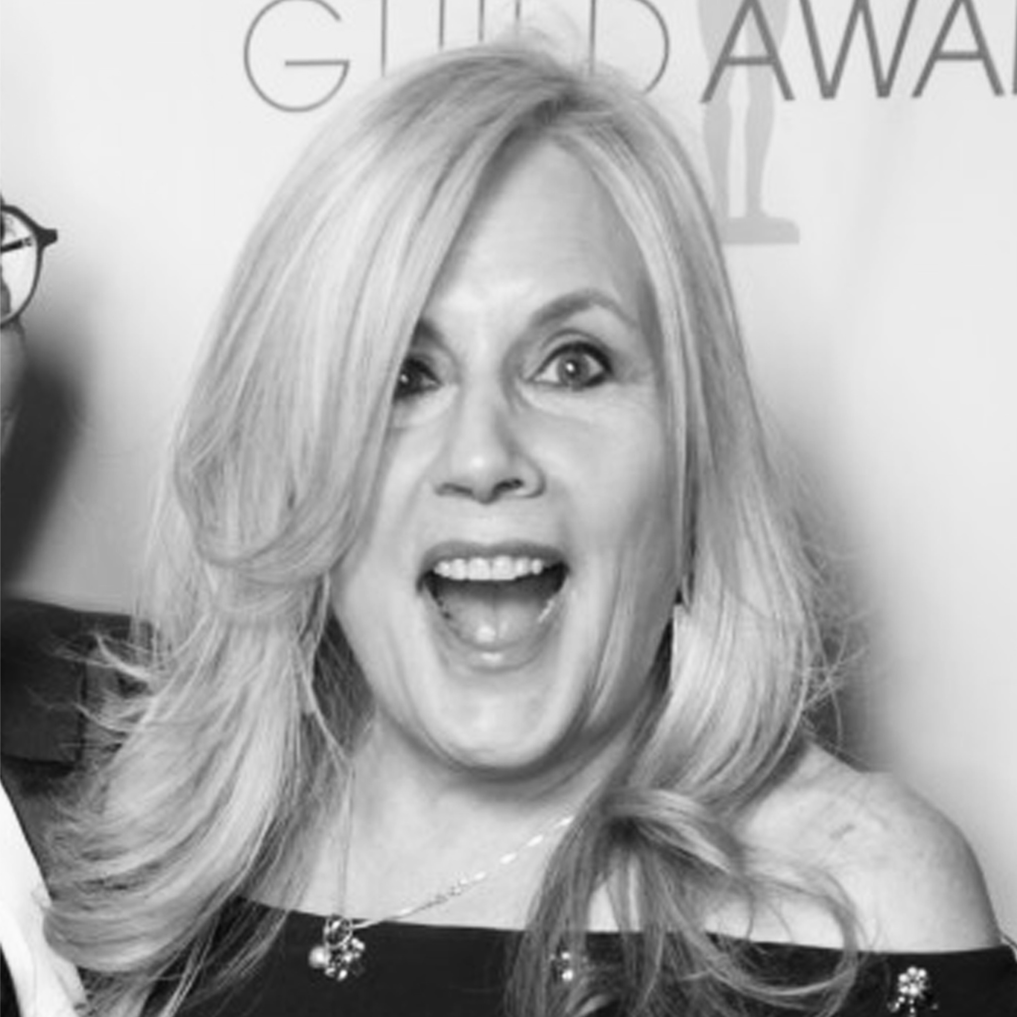 A woman with long blonde hair and expressive eyes smiles broadly with her mouth open in excitement. She is dressed in an off-the-shoulder outfit and stands in front of a backdrop with the word 
