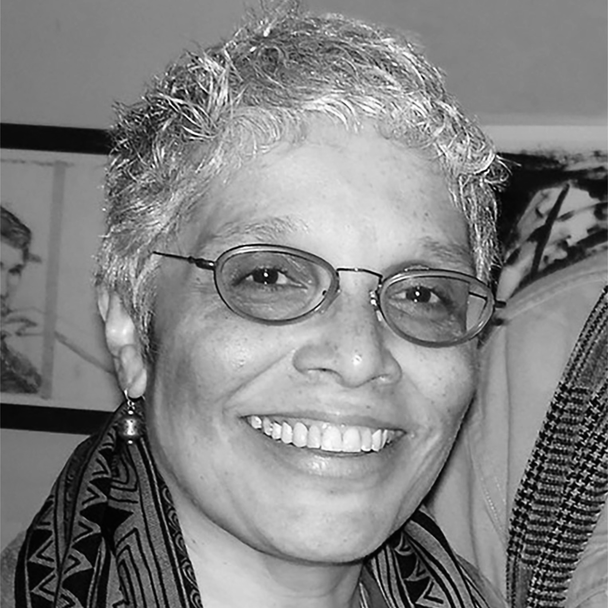An older woman with short, curly gray hair and glasses smiles warmly at the camera. She is wearing earrings and a patterned scarf. Behind her, part of a framed artwork is visible. The photo is in black and white.