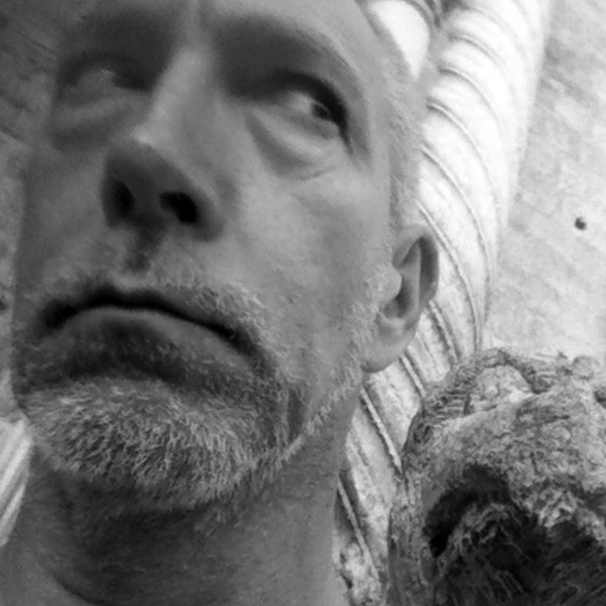 A close-up photo of a man’s face turned slightly, with a skeptical expression and looking up to his left. He has a trimmed beard and stands near a sculptural column, creating an artistic and candid black and white composition.