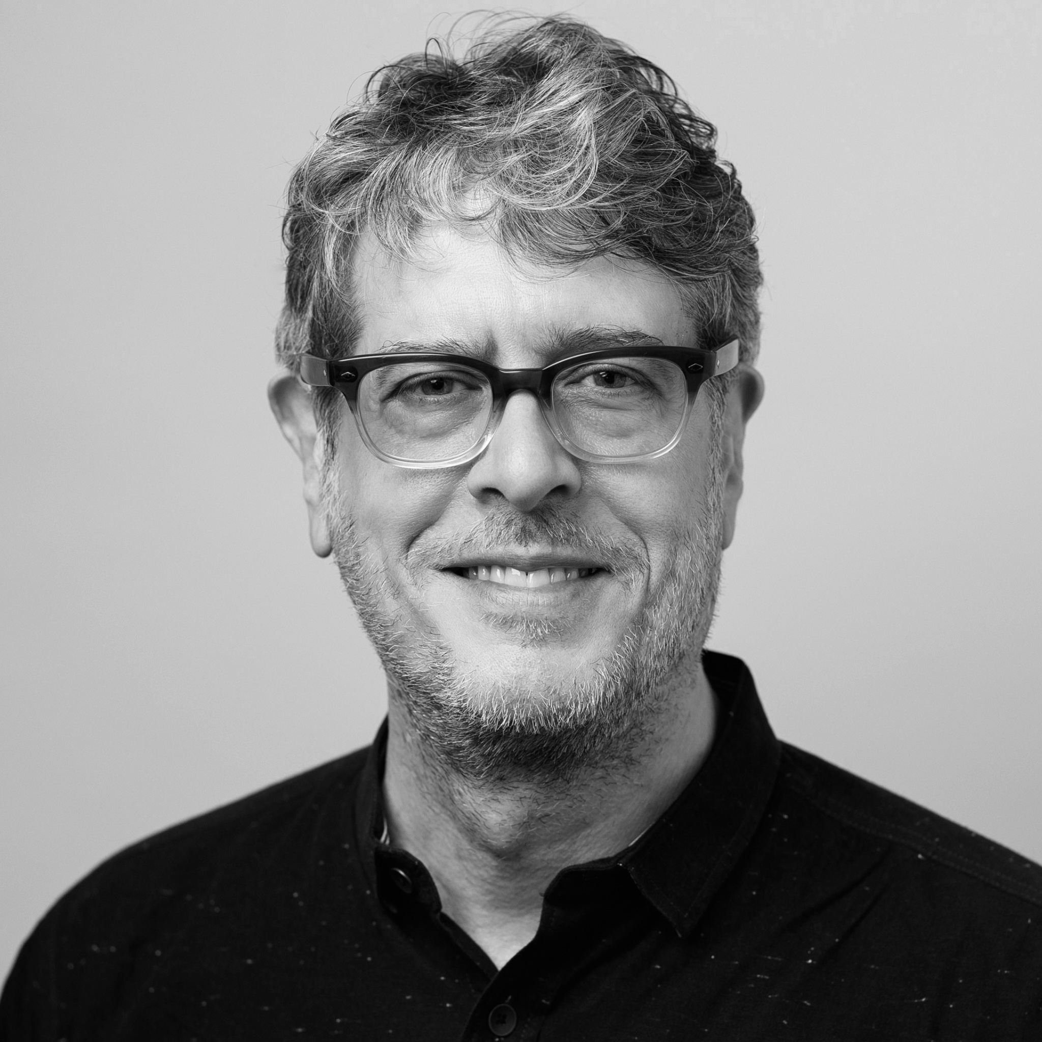 A man with slightly tousled gray hair and glasses smiles warmly at the camera. He wears a dark button-up shirt, and the background is a neutral studio tone. The image is in black and white with even lighting.