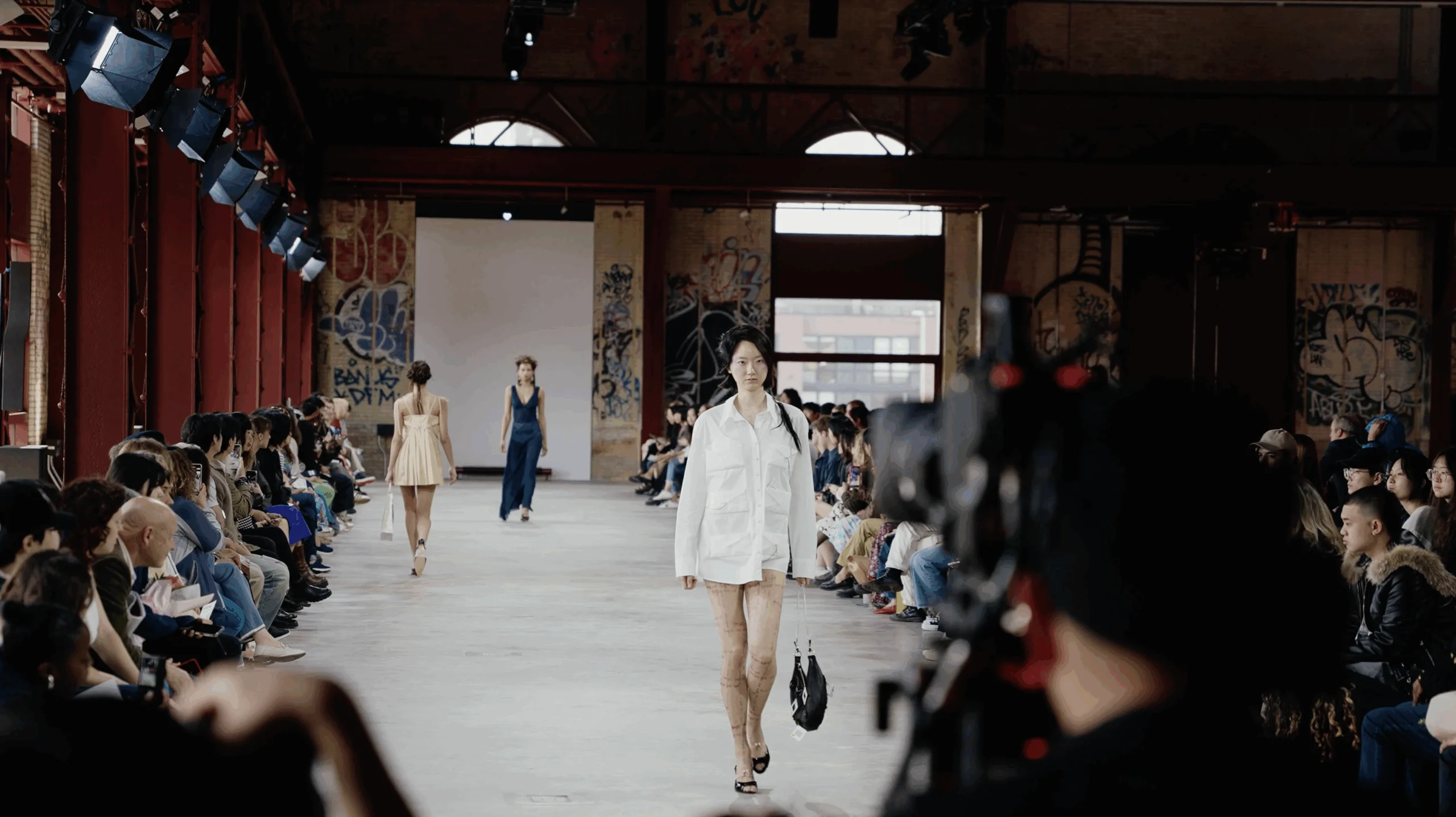 A fashion runway scene shows models walking down a catwalk lined with an audience. One model is dressed in a white oversized shirt and shorts while carrying a small bag. Another model, seen from behind, wears a light-colored dress, and a third model in a dark dress walks further down the runway. The venue features high ceilings with visible graffiti on the walls, and the crowd is engaged, with some taking photos.