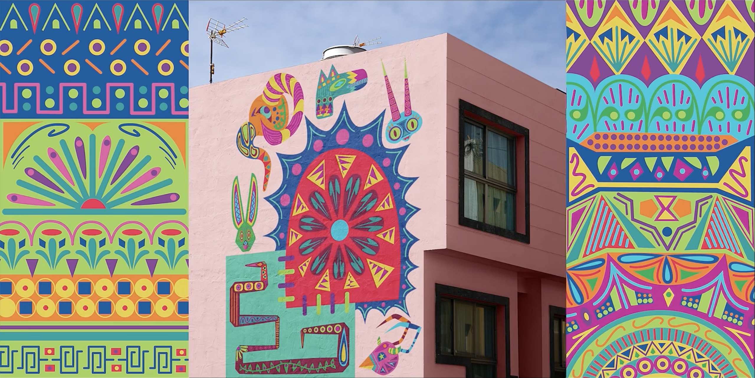 A vibrant mural covers the side of a pink building, featuring a central circular design with a blue circle surrounded by red and pink patterns. Colorful motifs, including animals and intricate shapes, adorn the mural, alongside decorative borders in various bright colors on the left and right sides. The sky above is clear, and a small antenna can be seen on the rooftop.