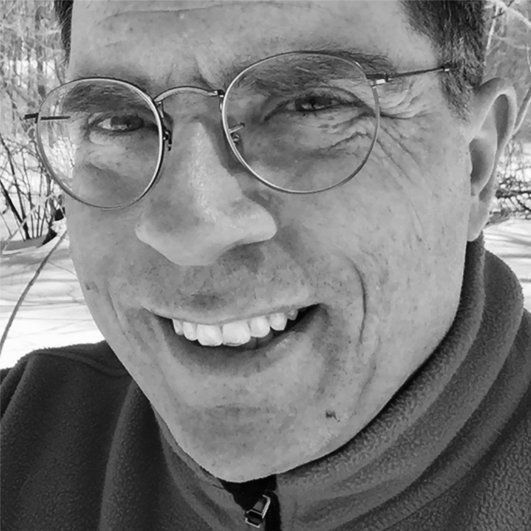 A man with round glasses smiles outdoors in the snow. He wears a fleece jacket and his face is partially in shadow. The close-up black and white image captures a cheerful winter moment.