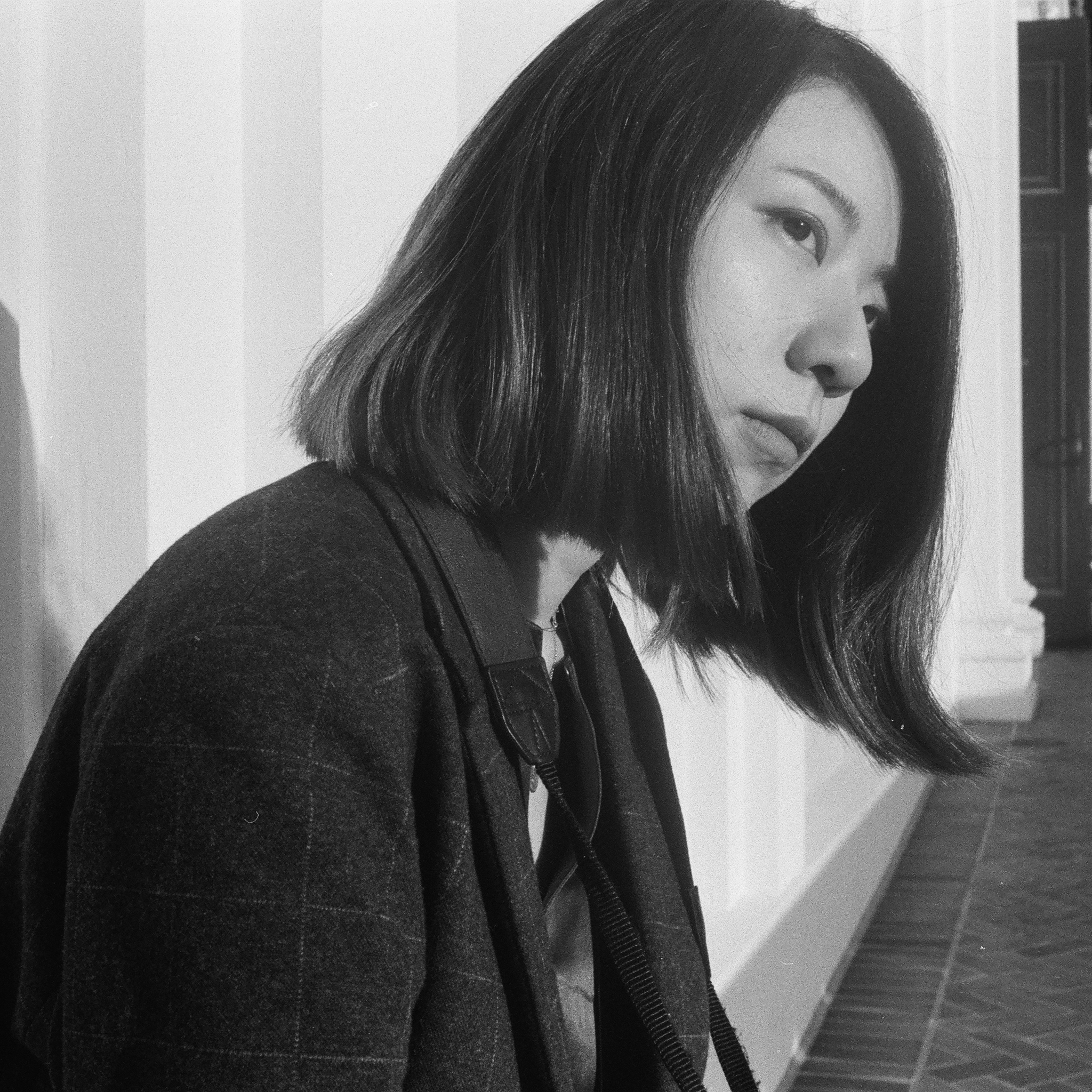 A woman with shoulder-length dark hair sits in profile, leaning slightly forward while gazing contemplatively into the distance. She wears a textured blazer and is lit by natural light streaming through tall, white columns. The background includes classical architectural elements and a patterned floor. The image is in black and white, with high contrast between light and shadow.