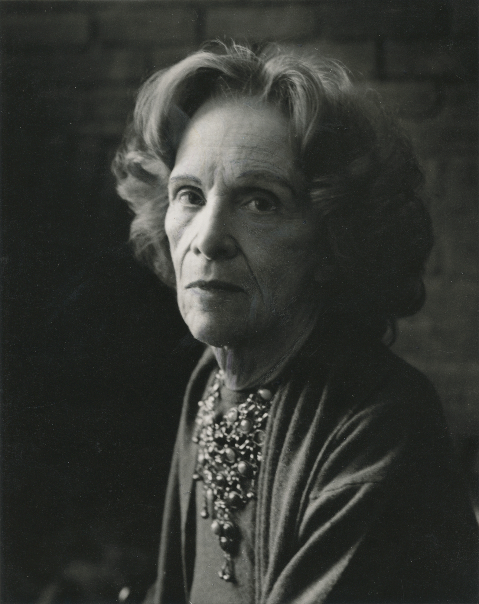a black and white photograph of an elderly white woman looking at the camera, wearing an elaborate necklace and a sweater