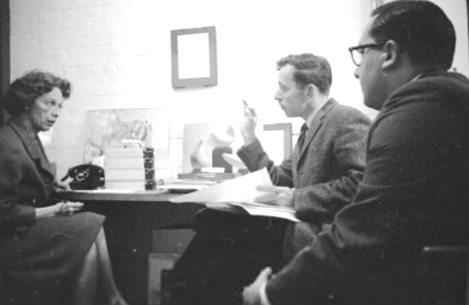 a black and white photograph of a white woman sitting on one side of a desk, talking to two white men on the other side of the desk. all are wearing mid-20th century attire