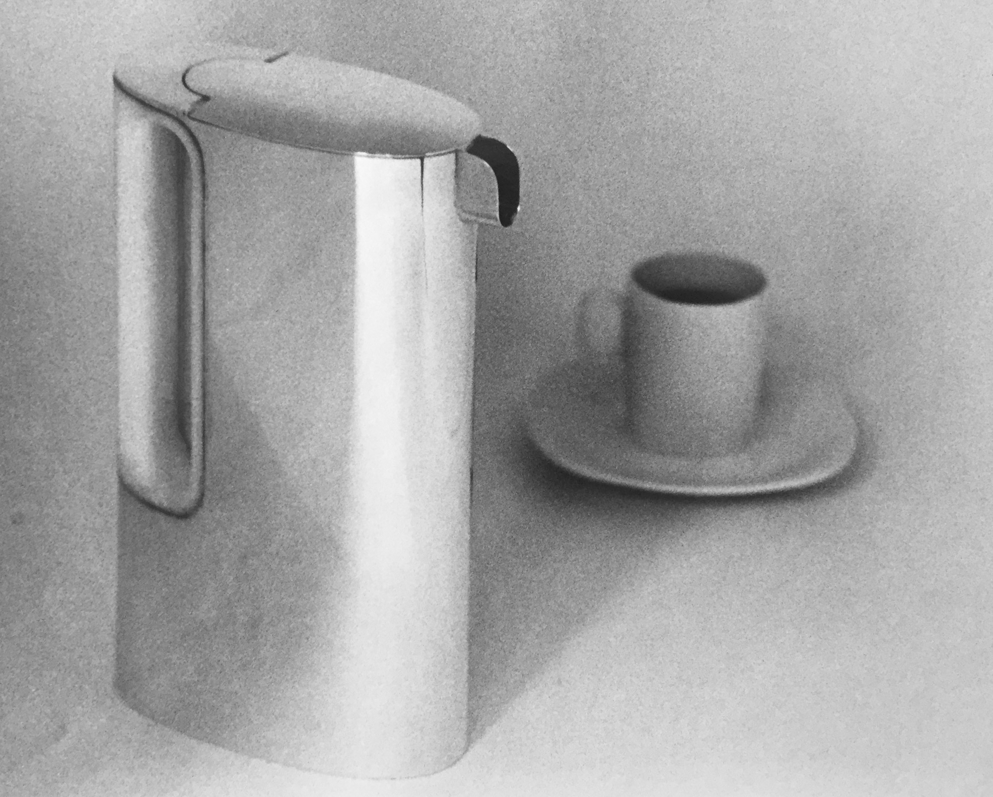 a black and white photograph of a stretched cylindrical coffee pot and a cup and saucer