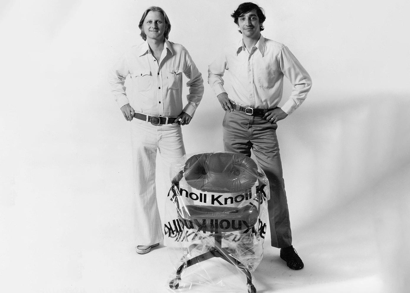 a black and white photograph of two white men with their hands on their hips. both are wearing white long sleeve button down shirts. one is wearing white pants, and the other jeans. they are standing behind a chair wrapped in plastic