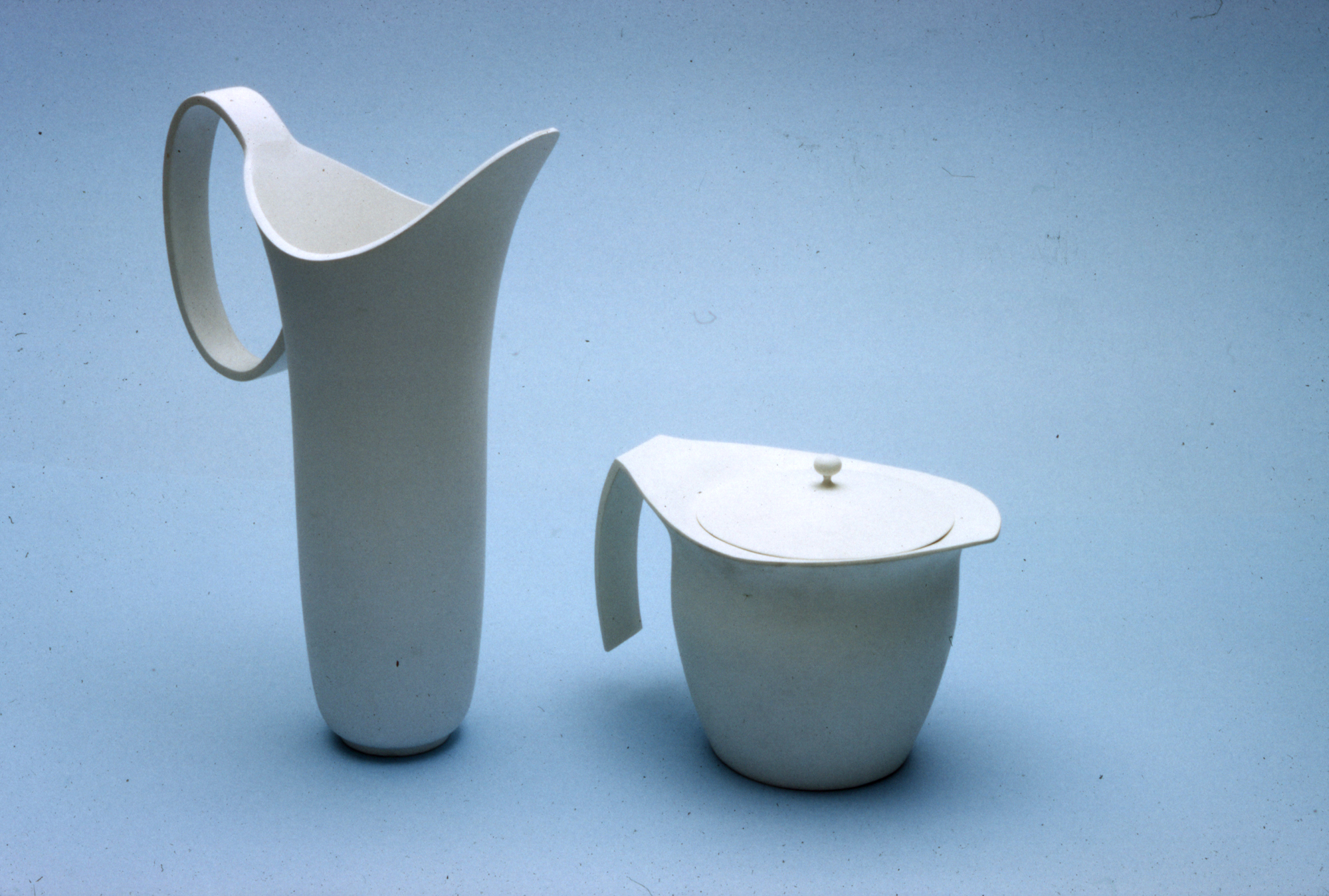 two white pitchers; one is tall and thin with a tapered handle and spout, the other is short and stout with a flat lid