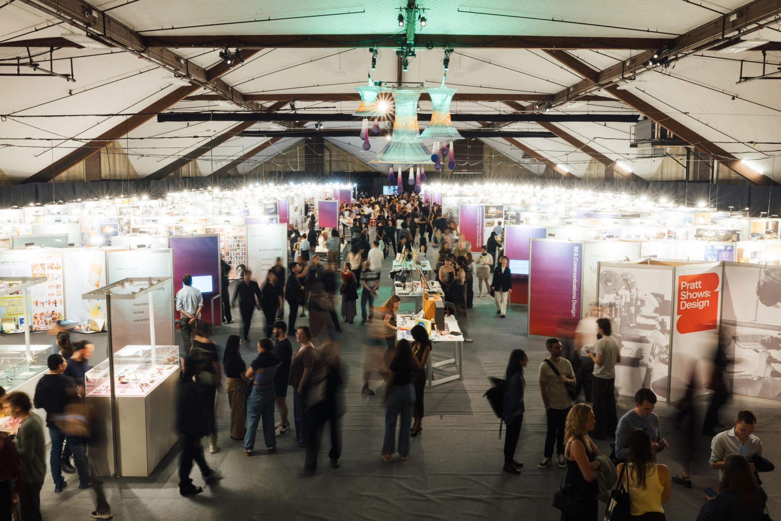 Wide overhead view of a crowded indoor design exhibition with rows of display booths and signage, as visitors walk through aisles beneath a high arched ceiling and hanging installation.
