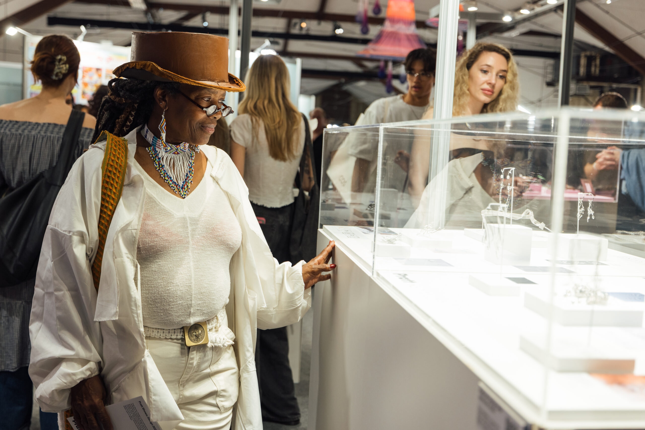 Visitor in a hat and layered white clothing views jewelry displayed in a glass case at a crowded indoor design exhibition, with other attendees and booths in the background.