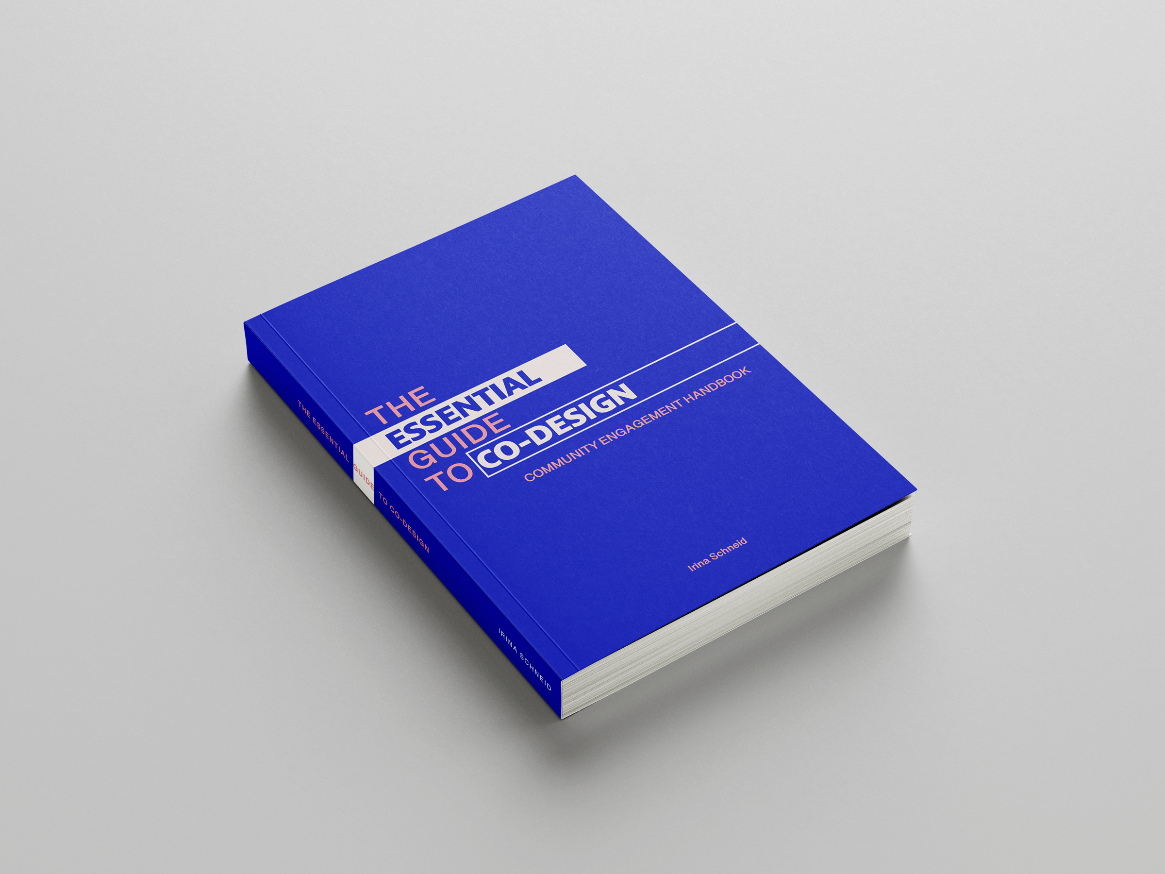 A book sits on a gray surface. The book is blue and is titled, The Essential Guide to Co-Design by Irina Schneid.