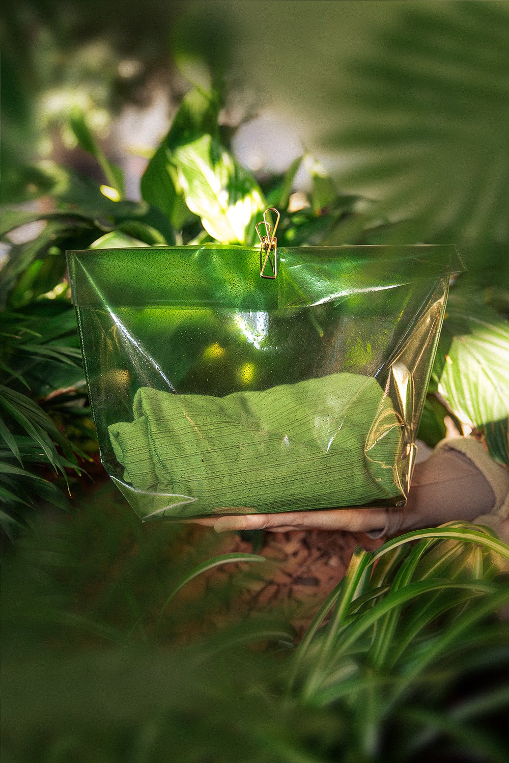 A hand holds a transparent green pouch, clipped at the top and photographed among greenery