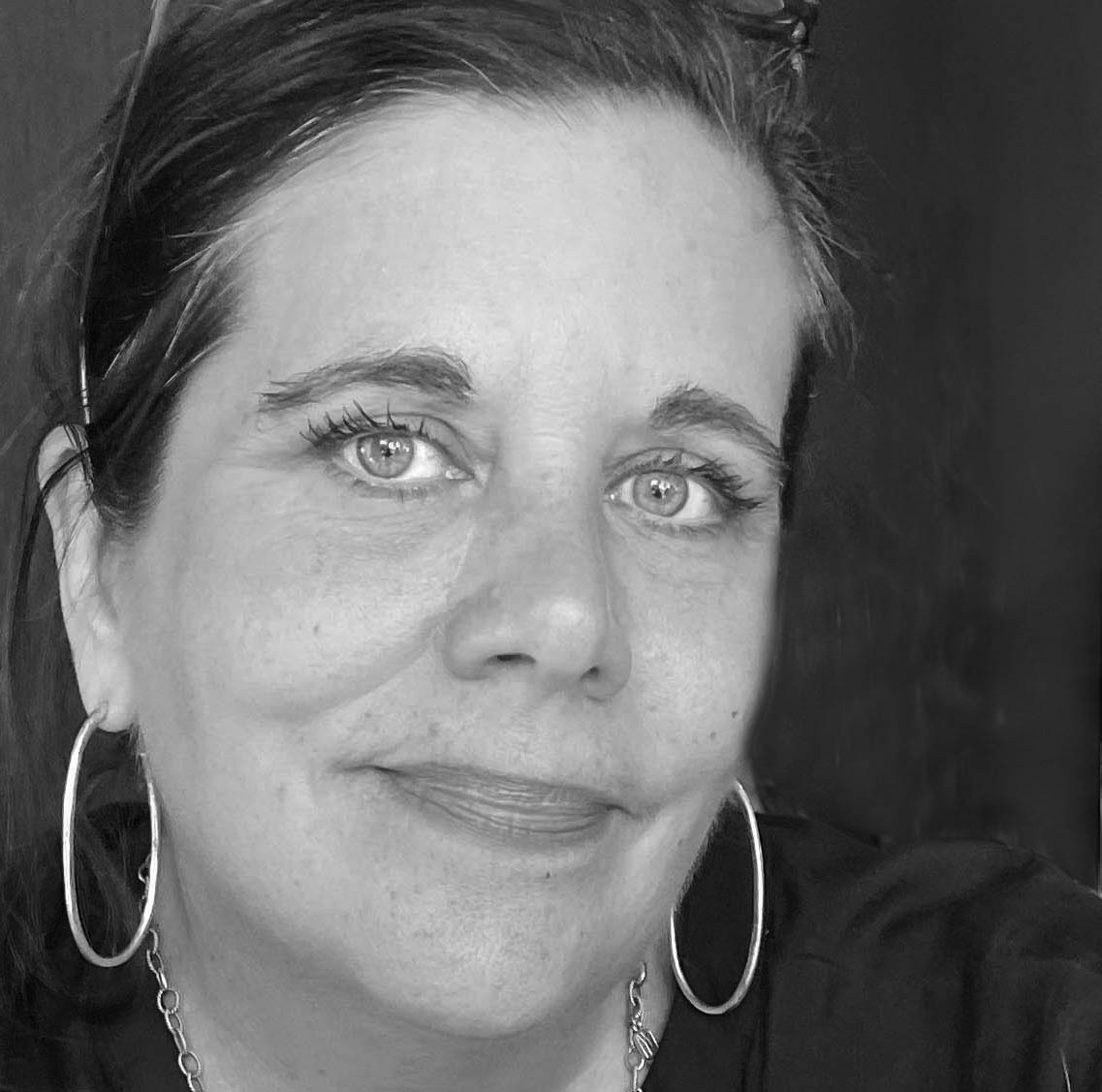 A close-up black and white portrait of a woman with light eyes and a relaxed smile. She wears large hoop earrings, a chain necklace, and has sunglasses resting on her head. Her hair is pulled back, and the background is dark and out of focus, drawing attention to her face.