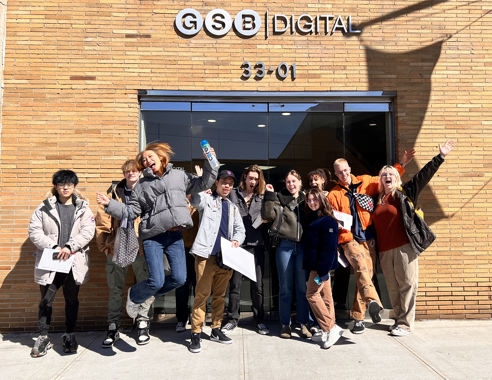A group of young people posing energetically in front of the GSB Digital building, with some smiling, jumping, and raising their arms in excitement. They are dressed in warm winter clothing, carrying papers and bags.