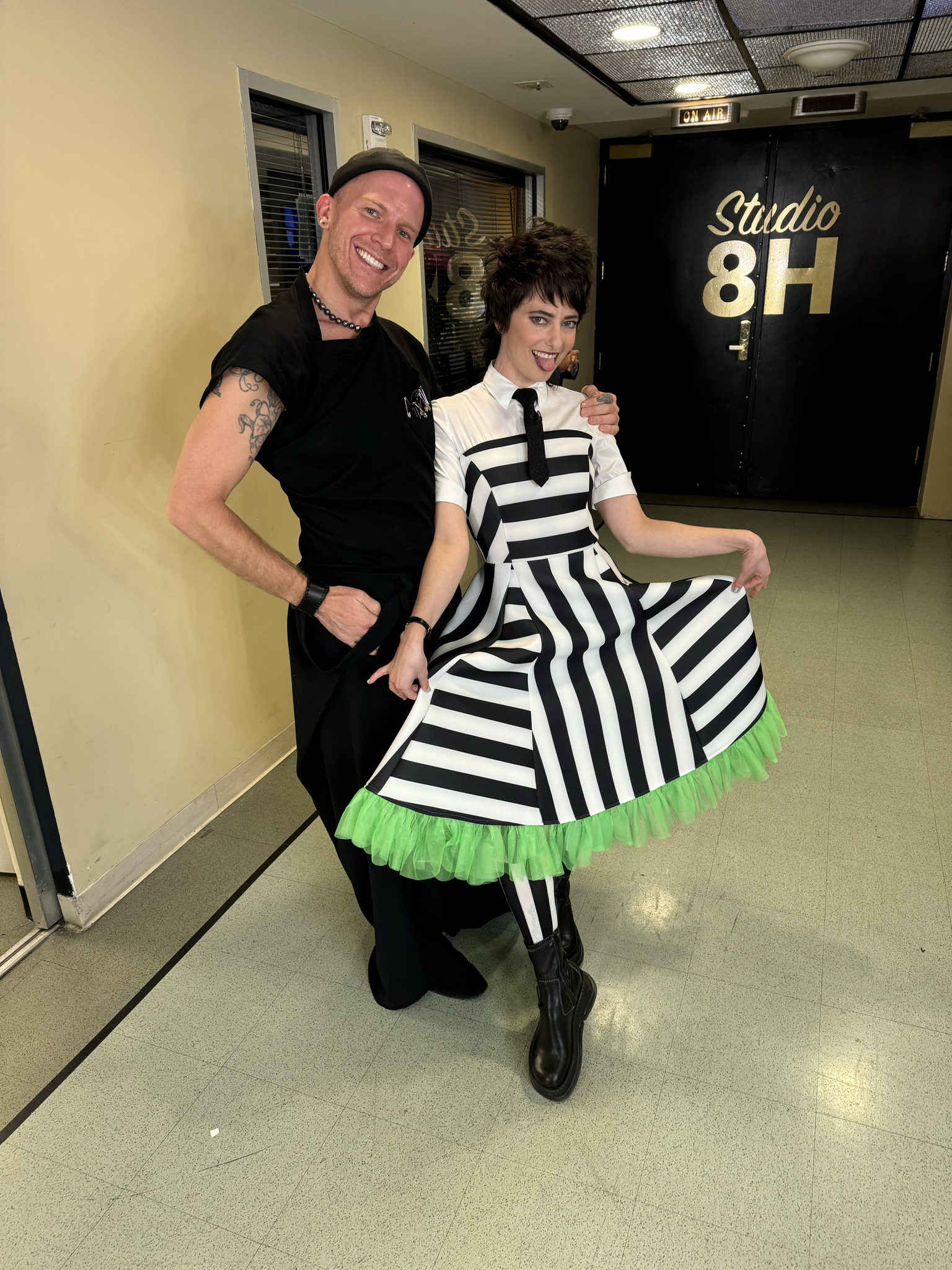 A cheerful designer and a performer pose in front of a “Studio 8H” sign. The performer is wearing a playful black-and-white striped dress with green tulle trim, styled like Beetlejuice, while the designer wears black and smiles broadly.