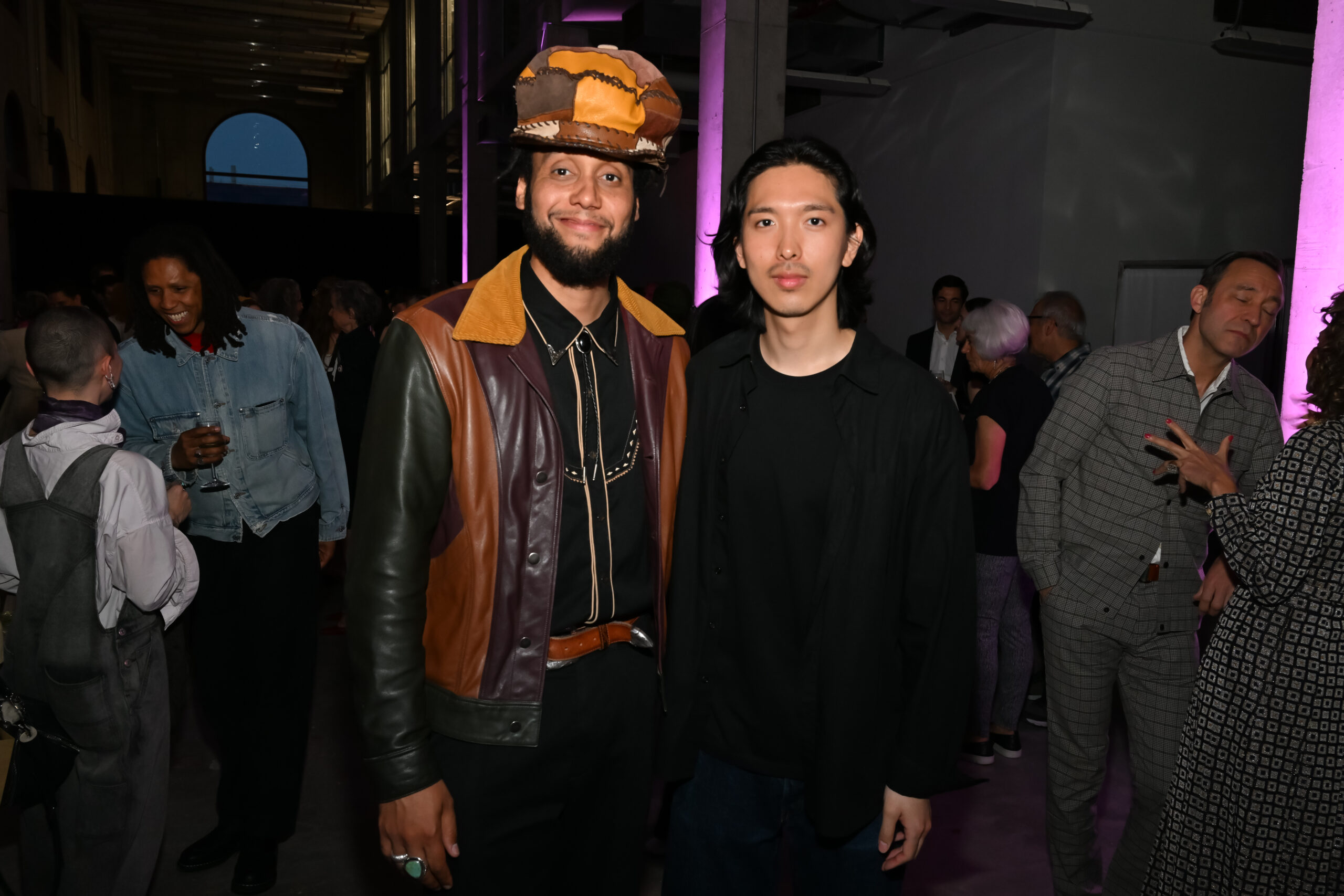 Two men pose together at a crowded indoor event, likely an art or fashion gathering. The man on the left wears a patchwork leather jacket in shades of brown, gold, and burgundy, with a bold oversized hat made of layered fabrics in warm tones. He smiles warmly and has a trimmed beard. The man on the right, dressed in all black with long dark hair, has a calm expression. The background is filled with people chatting under ambient purple lighting in a tall, gallery-like space with exposed beams and high ceilings.