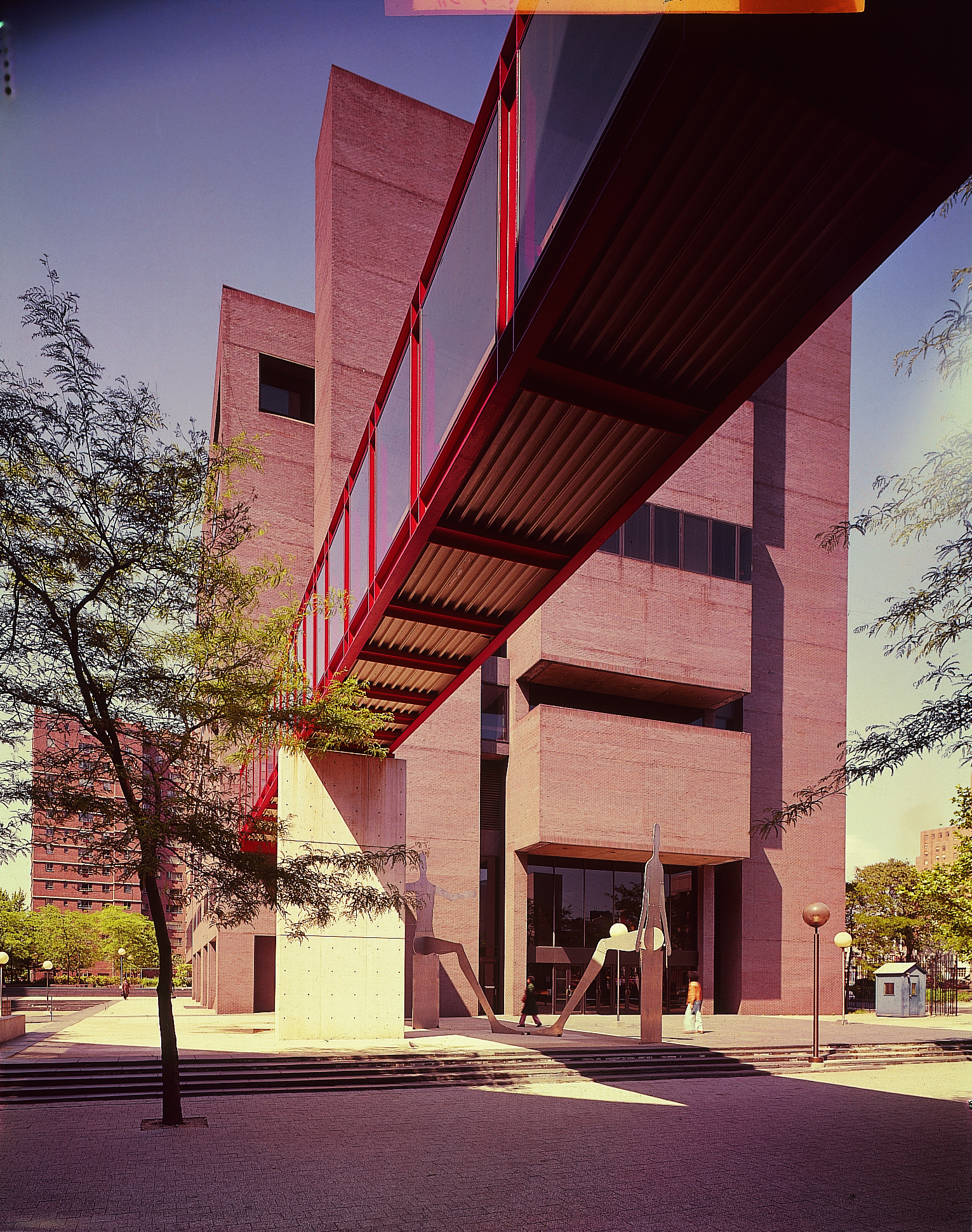 Davis Brody & Associates (Phyllis Birkby, project designer and co-project architect), exterior, Long Island University’s Library-Learning Center, Brooklyn, N.Y., 1975. Courtesy of Davis Brody Bond. Image displays a gray brutalist building with a red skybridge, with a single tree and metal sculpture (depicting a human form) in the foreground.