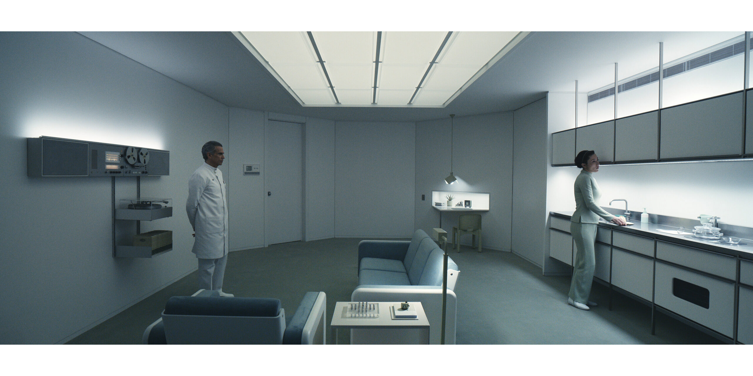 A futuristic, minimalist room with sterile white walls and muted furniture. On the left, a man in a white lab coat stands beside a wall-mounted reel-to-reel tape recorder and equipment. On the right, a woman in pale green clothing stands at a sleek counter with stacked glass dishes under bright lighting. In the center are modern blue sofas and a table with a chess set. The ceiling has a large illuminated panel, giving the room an austere, clinical atmosphere. The overall design feels retro-futuristic, reminiscent of mid-20th-century visions of space-age interiors.
