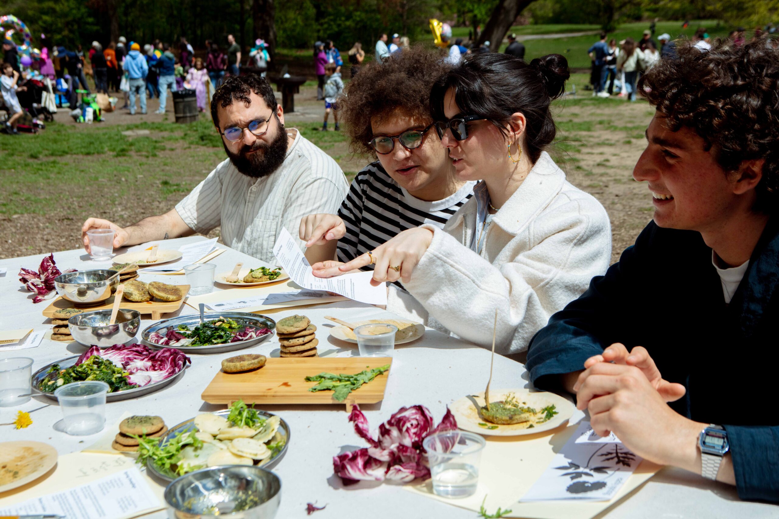 Four people sitting outdoors at a table covered with plates of greens and foraged food, engaged in conversation and looking over printed materials, with a crowd in the background.