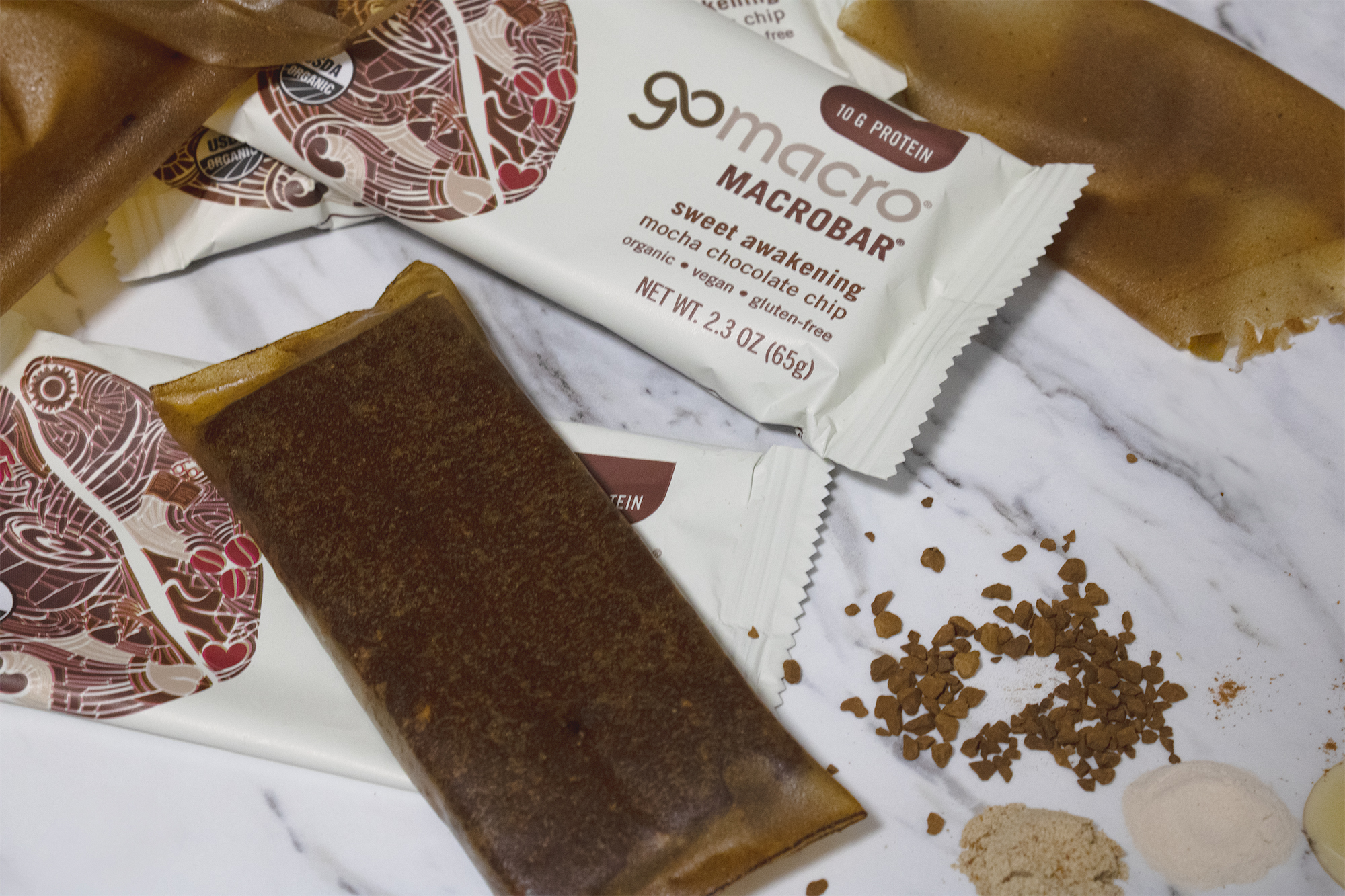 The image features several GoMacro Macrobars, specifically the "Sweet Awakening" flavor, displayed on a marble surface. Some bars are unwrapped and laying next to the packaged ones. Also present are scattered coffee granules and various powdered ingredients. The packaging prominently displays the brand name and product details, highlighting attributes like being organic, vegan, and gluten-free.
