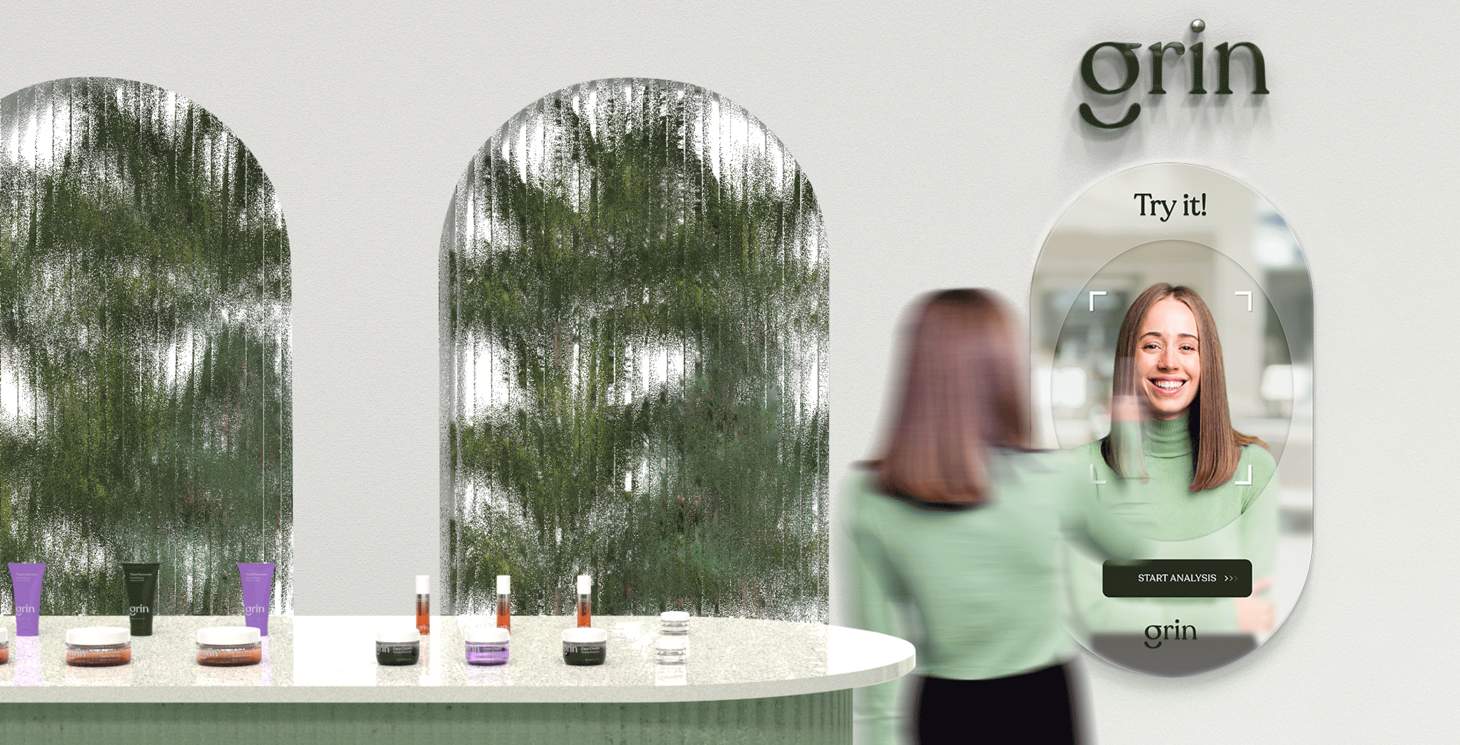 A person stands in front of a digital mirror displaying a facial analysis interface labeled "Try it!" and "START ANALYSIS." The individual is smiling and wearing a green sweater. In the foreground, there's a countertop with various skincare products in different containers and tubes, featuring the brand "grin." The background features arches with a blurred green backdrop resembling foliage.