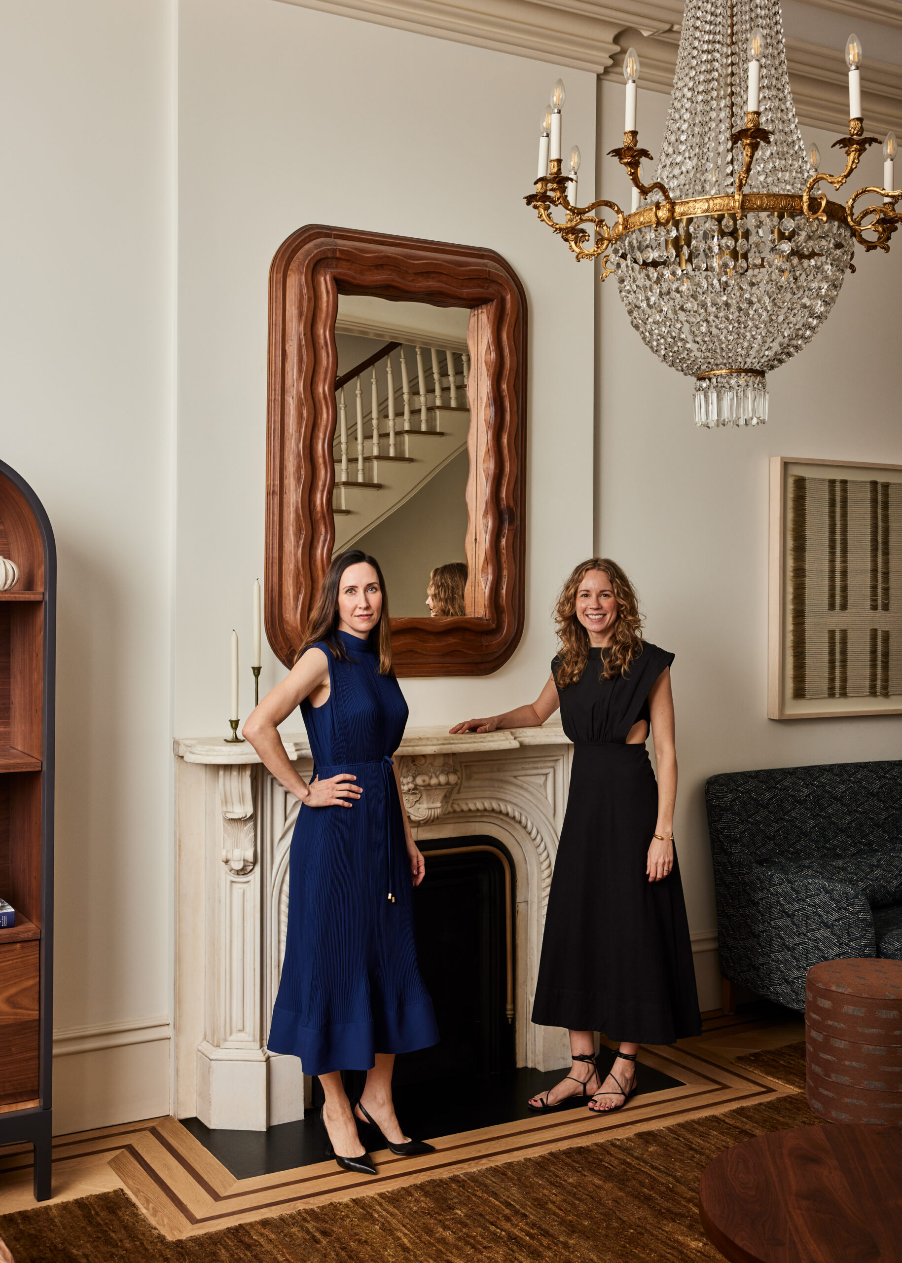 Amanda Jesse and Whitney Parris-Lamb stand in front of a historic fireplace. Two candlesticks rest on the mantle. A mirror with a wavy oak frame hangs above the fireplace. From the ceiling hangs a gold chandelier.