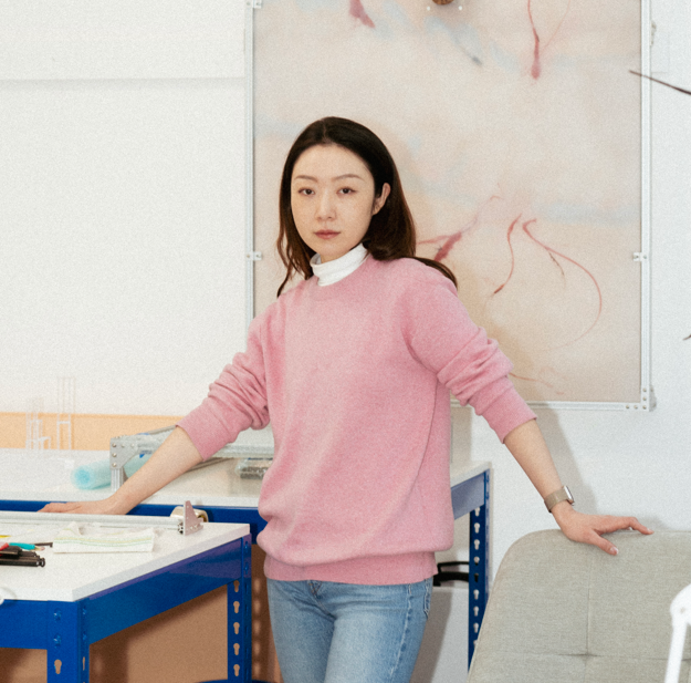 A woman stands in a modern studio workspace, leaning casually between a blue metal worktable and a gray chair. She is wearing a light pink sweater layered over a white turtleneck and light blue jeans, with a silver watch on her left wrist. Her expression is calm and focused, with a direct gaze toward the camera, conveying quiet confidence. The background features a clean, minimal workspace with white walls, scattered design tools, and a large abstract artwork on the wall behind her—pale beige with soft red and pink organic lines that resemble veins or flowing ink. The lighting is natural and evenly diffused, giving the image a serene, contemplative tone. This photograph is likely a portrait of an artist or designer in her studio environment, emphasizing clarity, simplicity, and an understated aesthetic that aligns with creative professionalism.