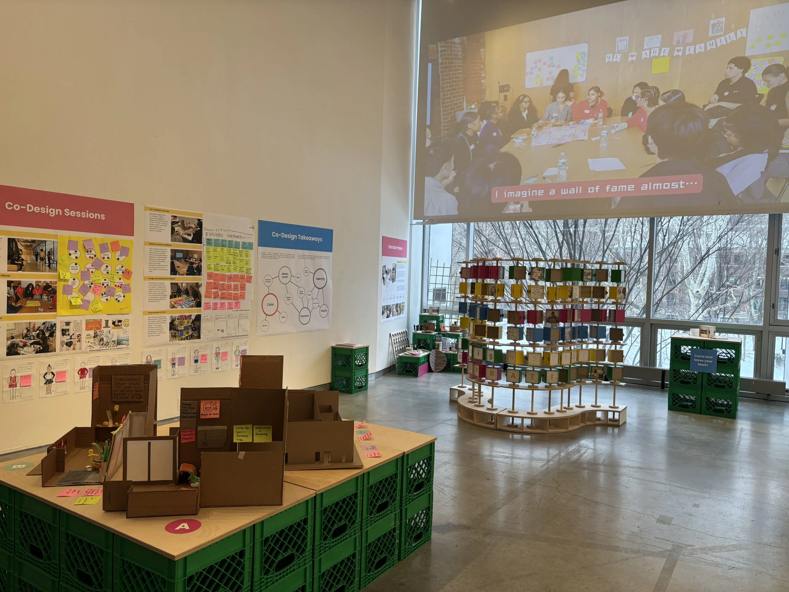 Wide view of an exhibition gallery space featuring a participatory design installation. In the foreground, cardboard architectural models and materials are arranged on a table supported by green plastic crates. The walls display posters, diagrams, sketches, and sticky notes labeled with headings such as “Co-Design Sessions” and “Co-Design Takeaways.” A large projection on the wall shows a group discussion around a table. The space is well-lit with polished concrete floors and large windows along one side.