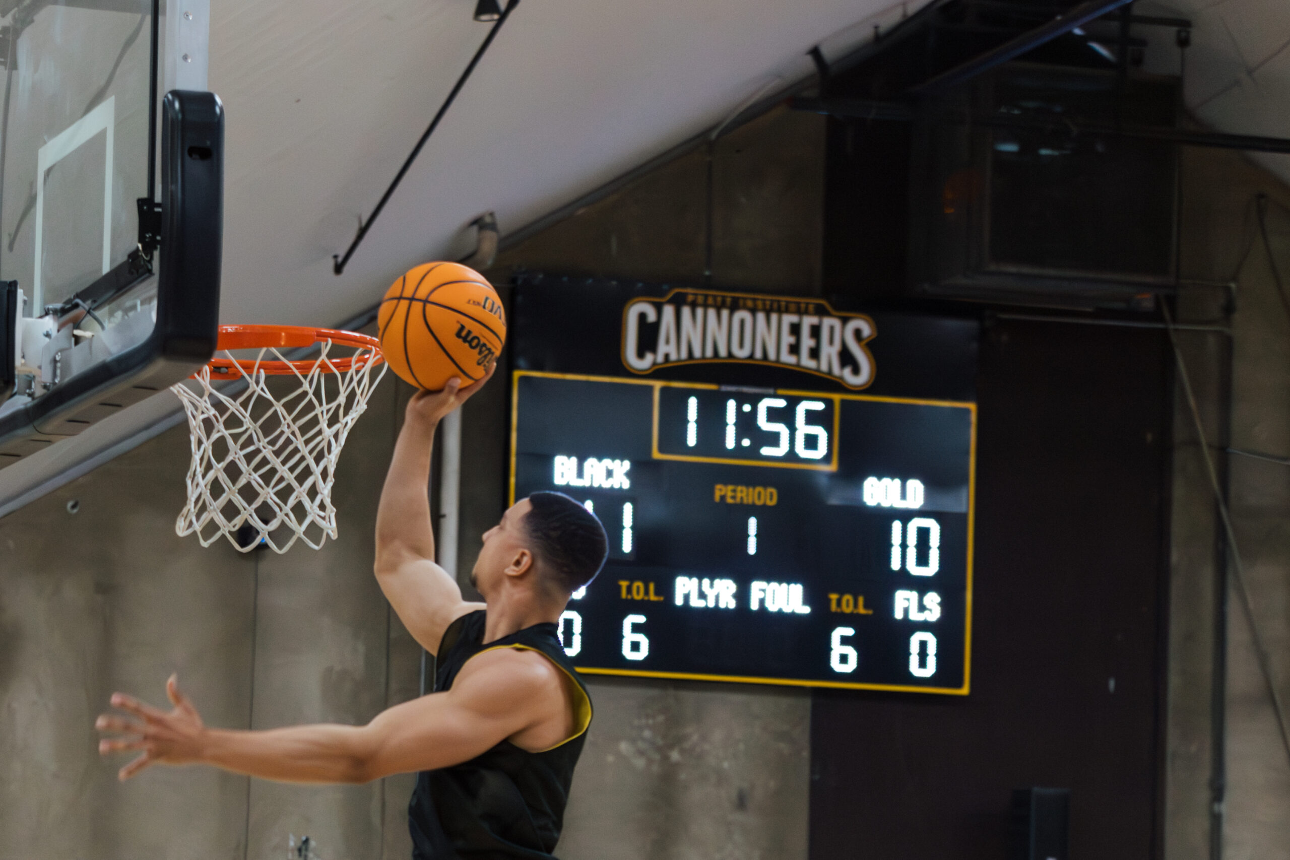 A basketball player is leaping towards the hoop to score a basket, holding a basketball in one hand. In the background, a scoreboard displays the time as 11:56, with a score of 1 for the black team and 10 for the gold team. The scoreboard also shows the current period and foul counts. The setting appears to be an indoor basketball court.