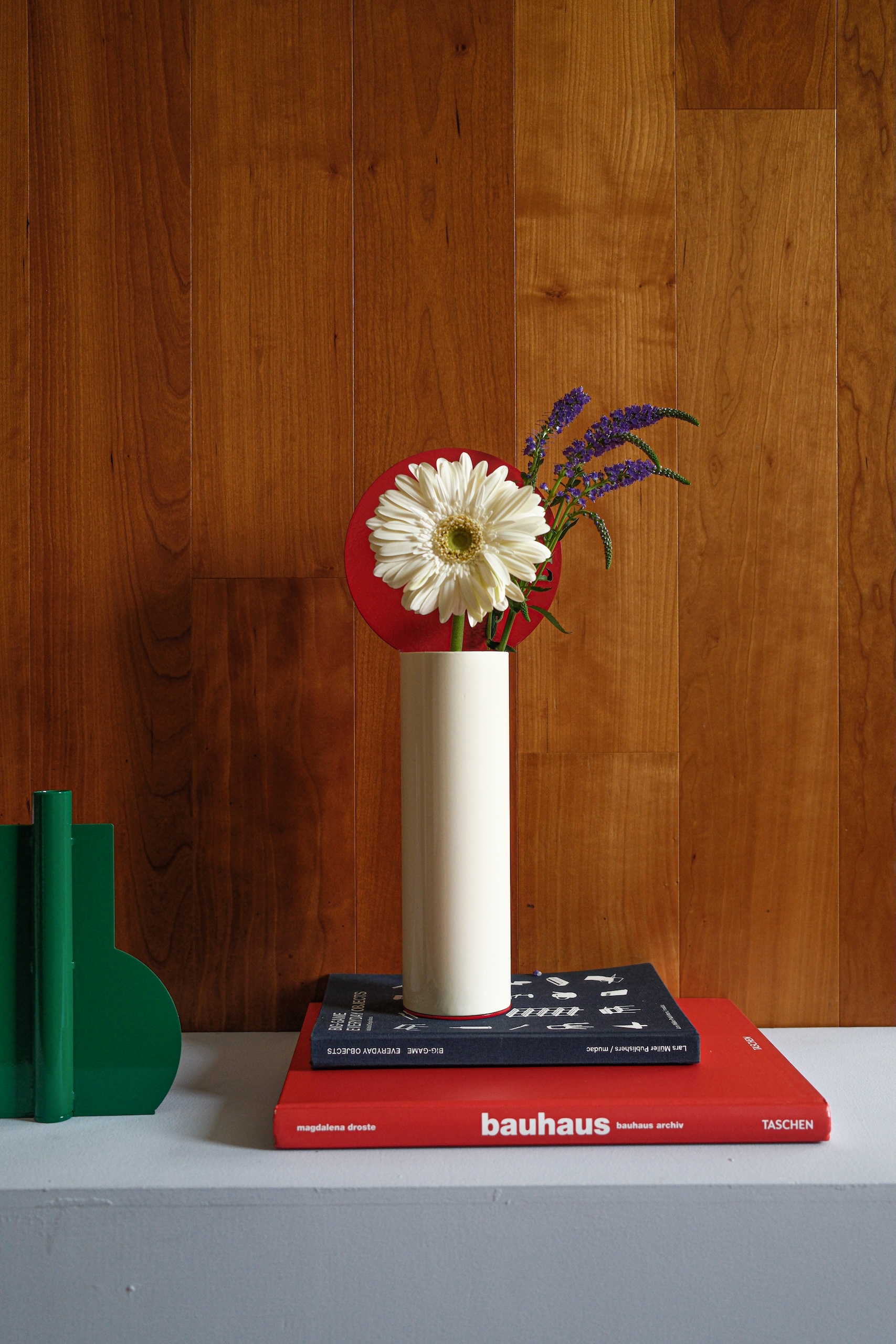 A vase displayed on a stack of design books holds a white Gerbera Daisy and several stalks of lavender. The vase has a thin white cylindrical form. Behind the white flower is a red disc form.